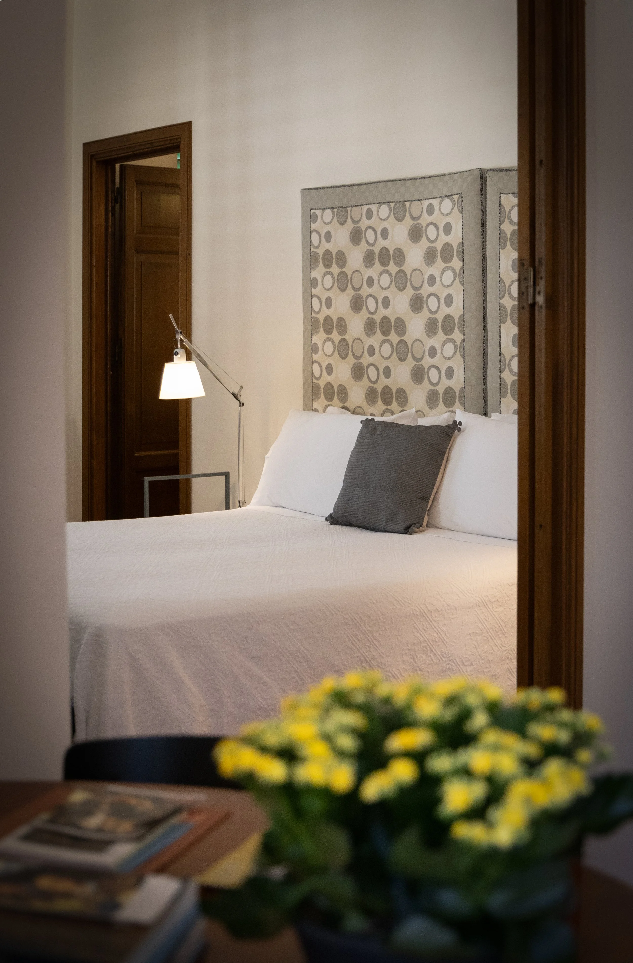 A hotel room viewed through a doorway, featuring a white bed with gray pillows, a patterned headboard, a wall-mounted adjustable lamp, and beside it a wooden door.