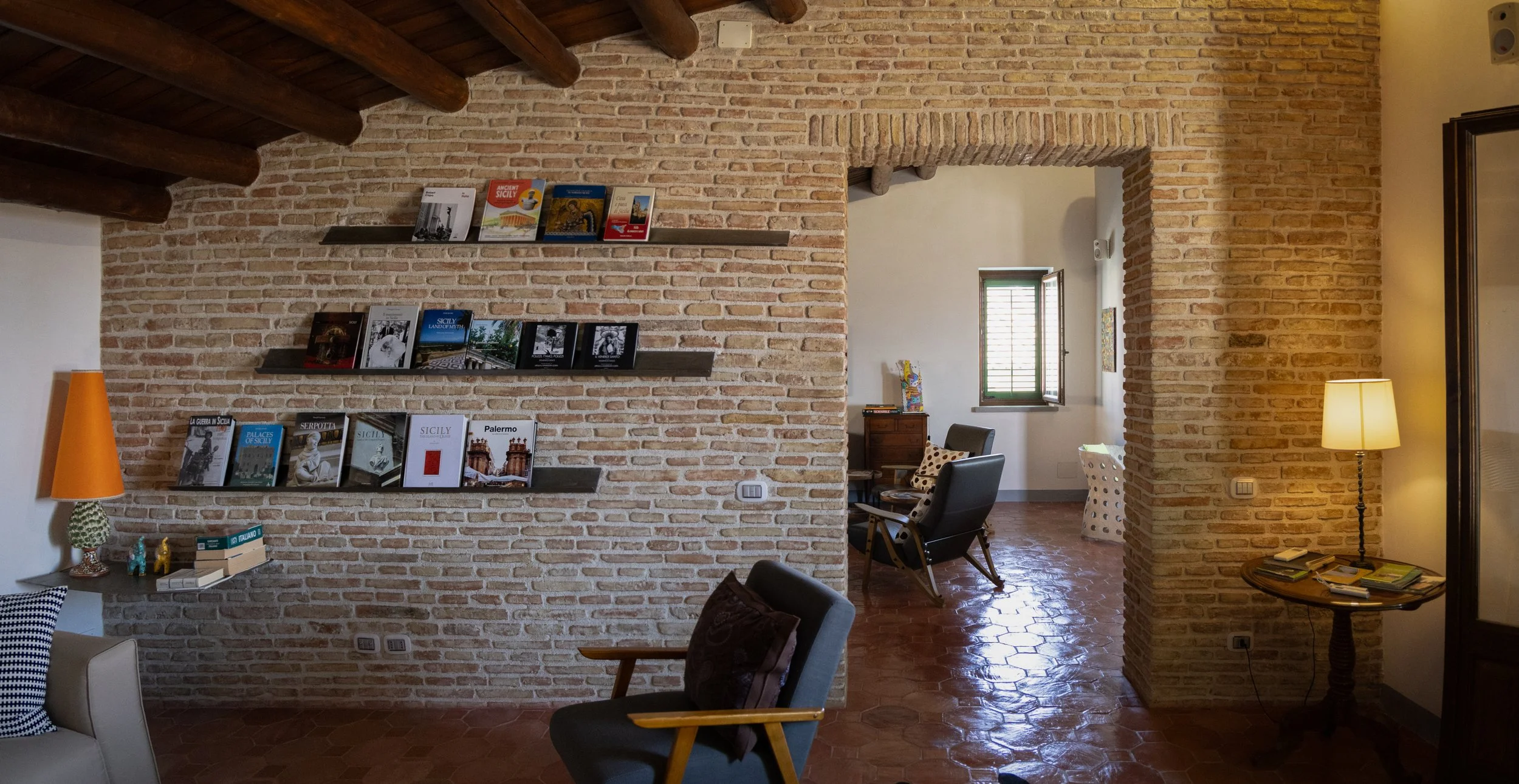 Living room with brick walls, wooden ceiling beams, bookshelves, armchair, lamps, window, and a small table with books.