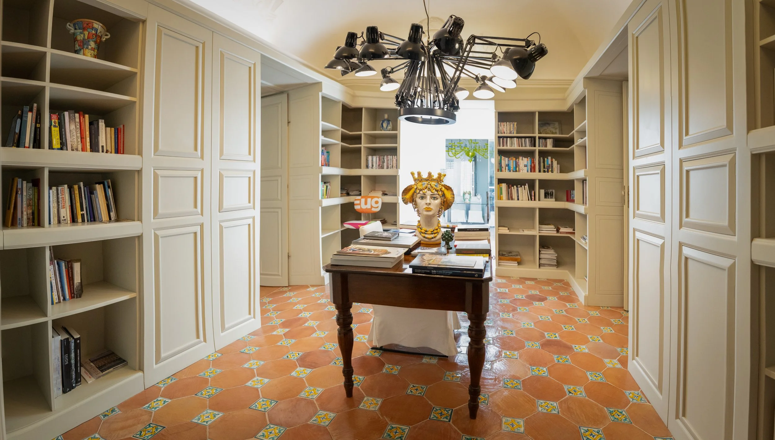 A room with white built-in bookshelves along the walls, a wooden table in the center with stacks of books, a decorated mannequin head with jewelry on the table, and a black chandelier light fixture hanging overhead. The room has a terracotta tiled fl