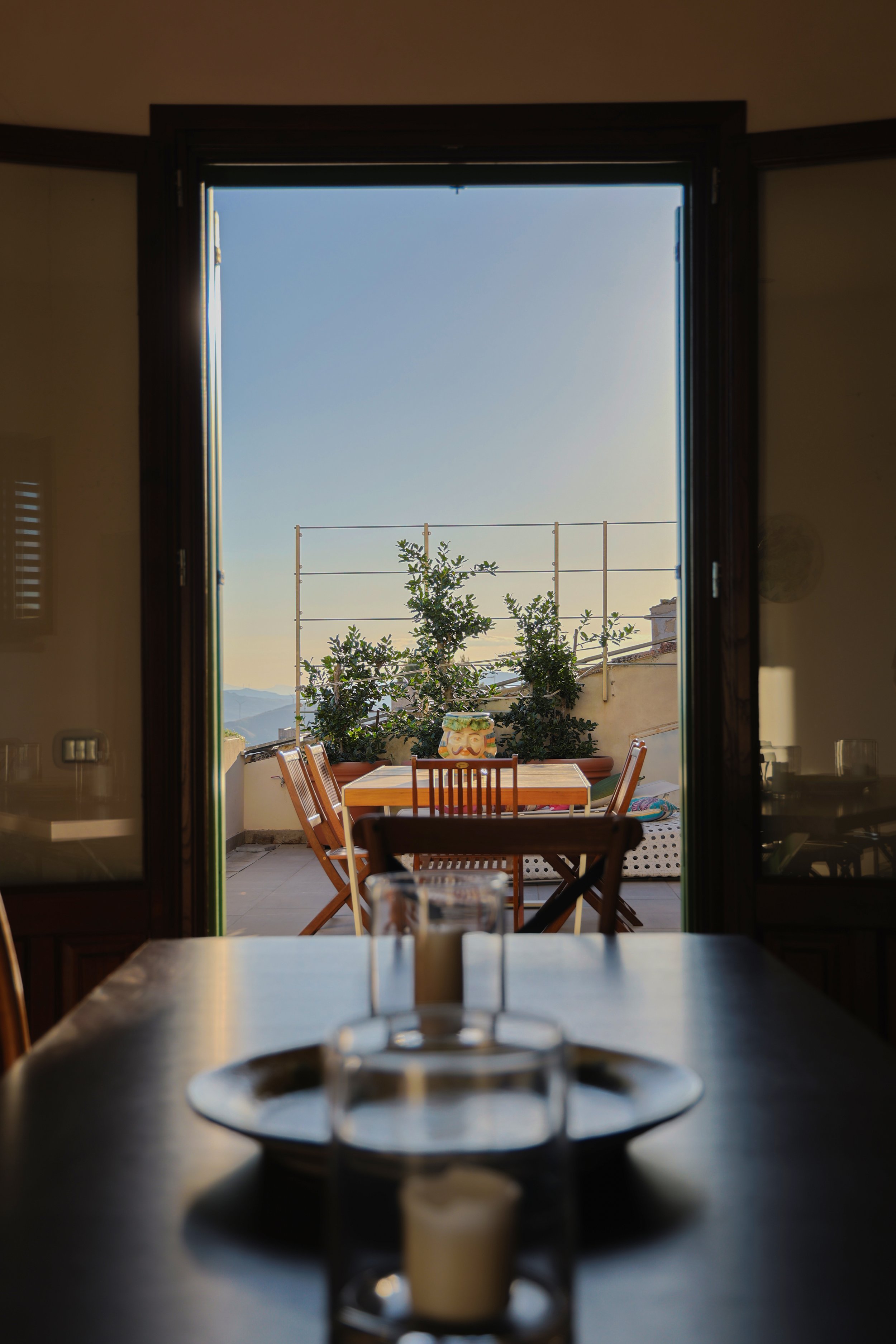 Interior view of a dining room looking out onto a balcony with outdoor furniture and plants under a clear sky.