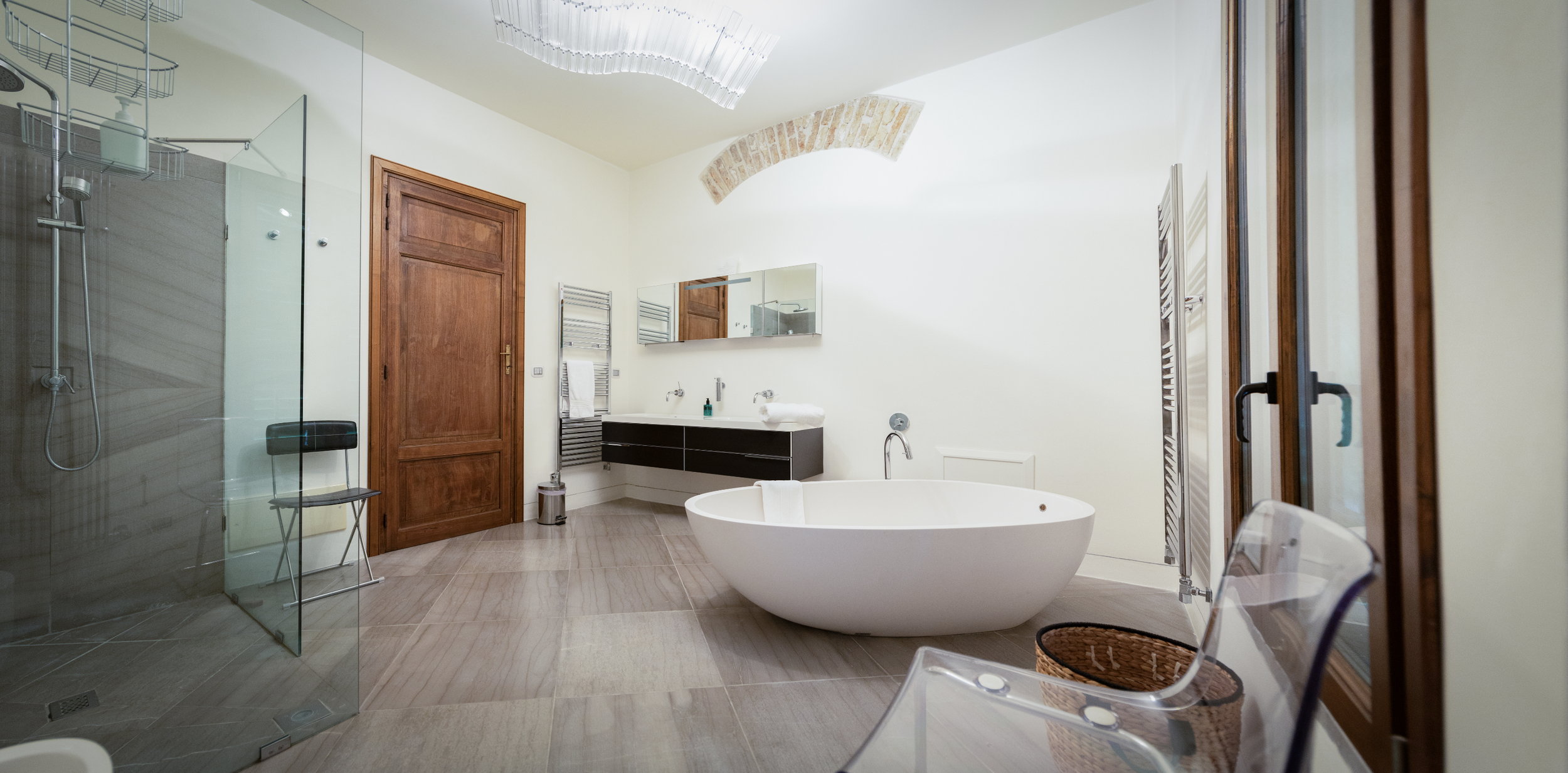 Modern bathroom with a freestanding bathtub, glass-enclosed shower, wooden door, wall-mounted sink with mirror, towel rack, wooden-framed windows, and wooden floors.