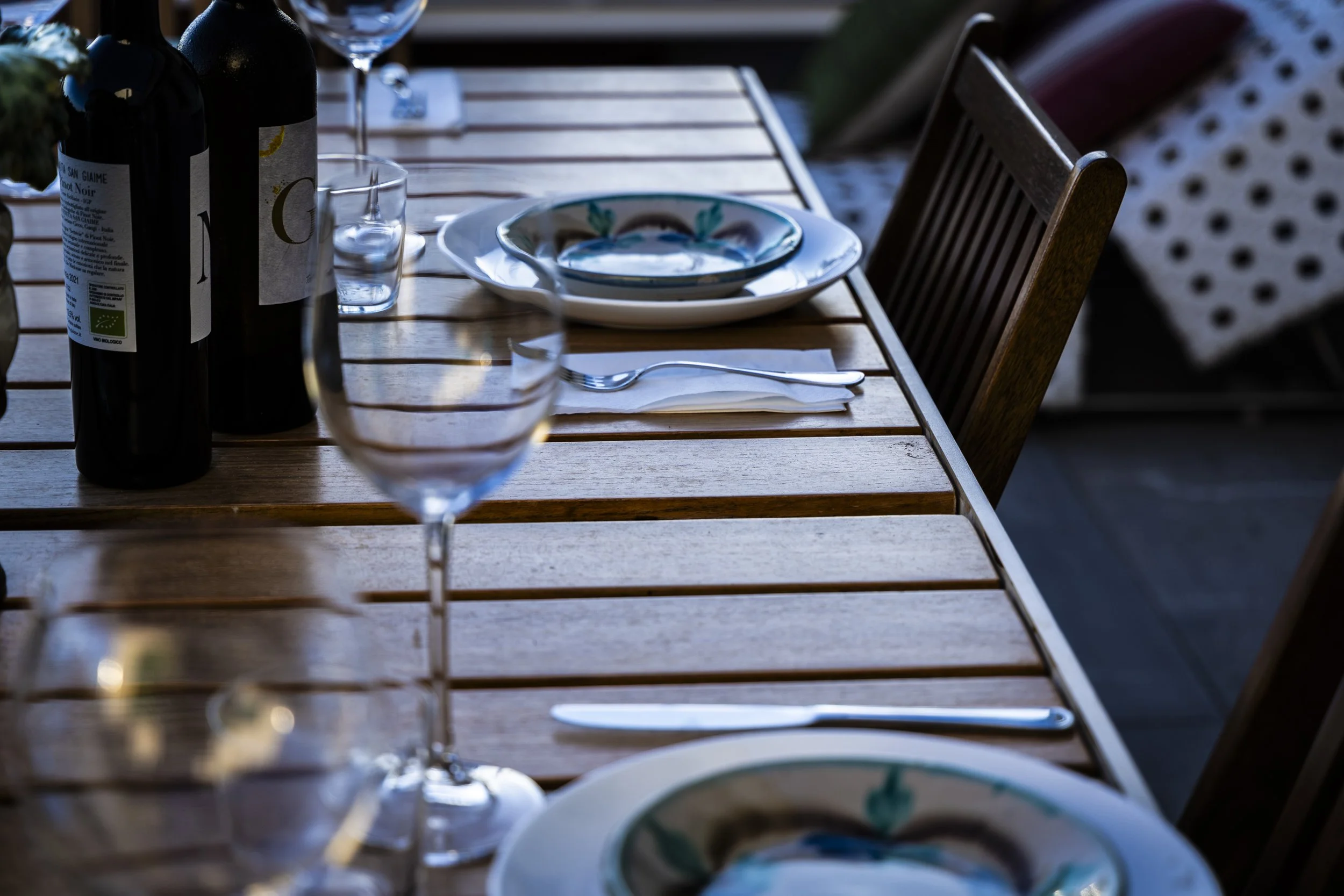 Empty wooden dining table set with plates, glasses, cutlery, and bottles of wine in an outdoor setting.