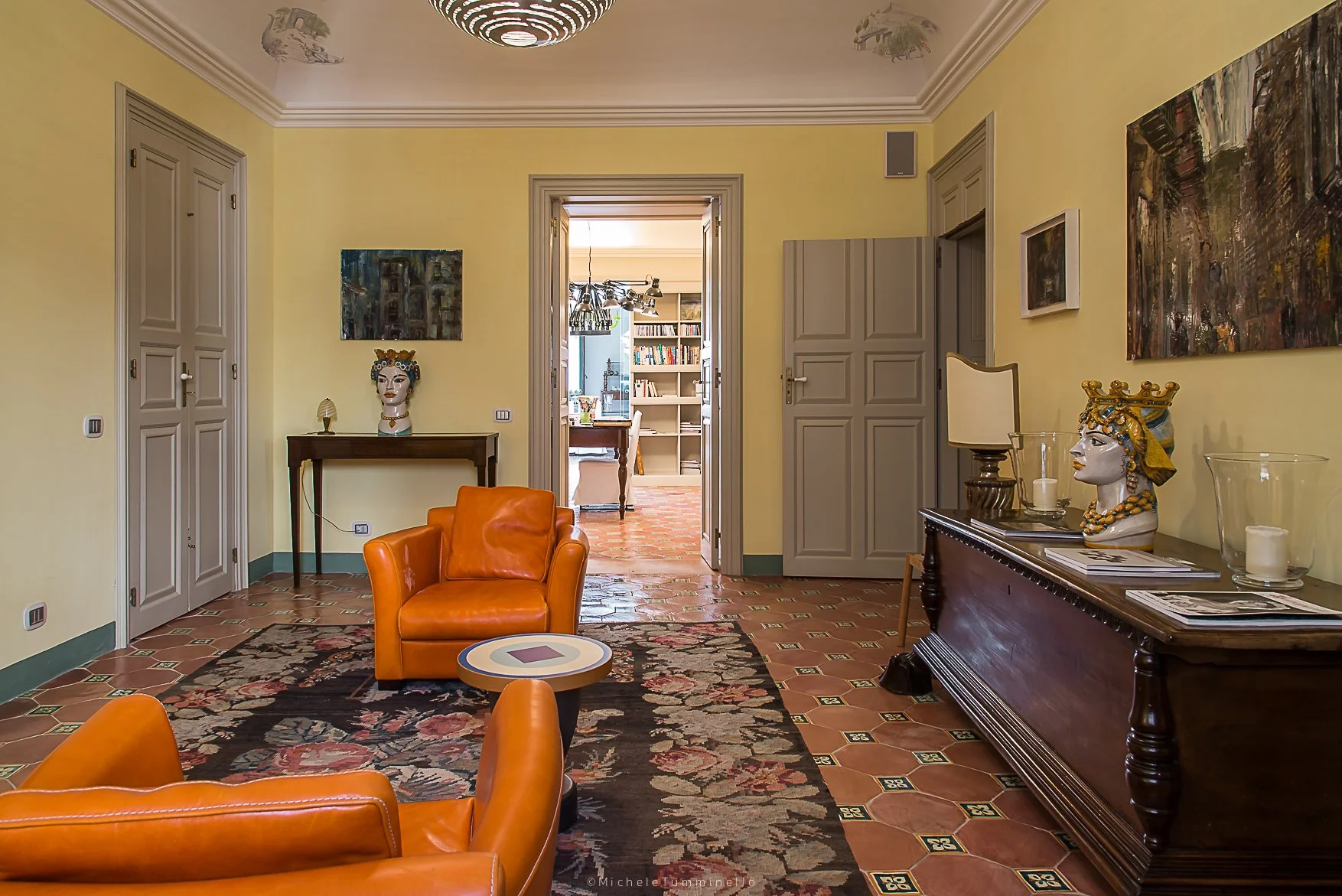 An interior living room with yellow walls, terracotta tiled floor, and a doorway leading into a study or library. There are two orange leather armchairs, a round coffee table, and several decorative busts and paintings. The ceiling has a modern circu