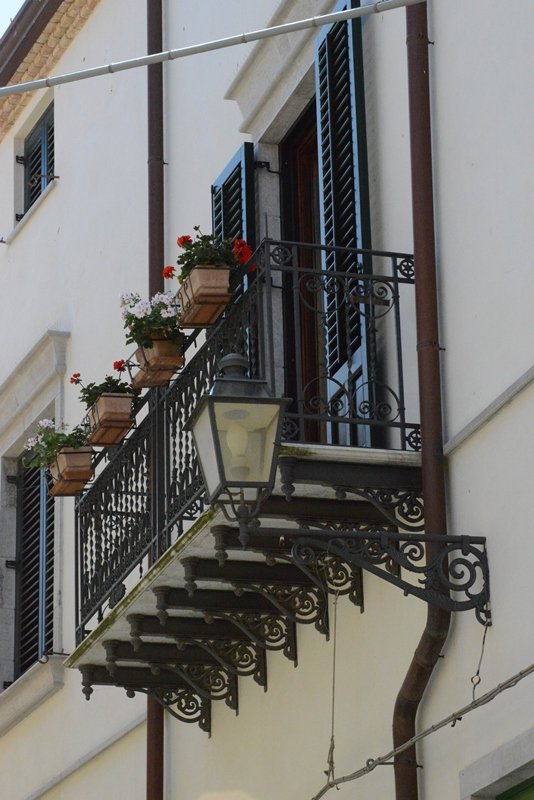 A balcony with decorative black wrought iron railing, potted flowers, a streetlamp, and blue window shutters on a white building wall.