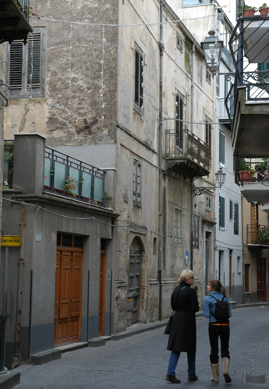 Two women walking and talking down a narrow, old European street with weathered buildings, balconies, and a sidewalk. One woman wears a long coat and glasses, the other has a backpack and boots.