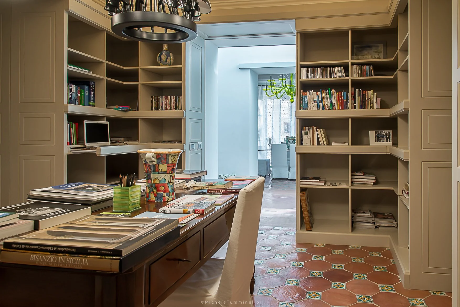 A home office with built-in bookshelves on either side filled with books and decorative items. A wooden desk with a beige chair is in the foreground, cluttered with magazines, books, and a colorful ceramic pot. The floor has patterned terracotta tile
