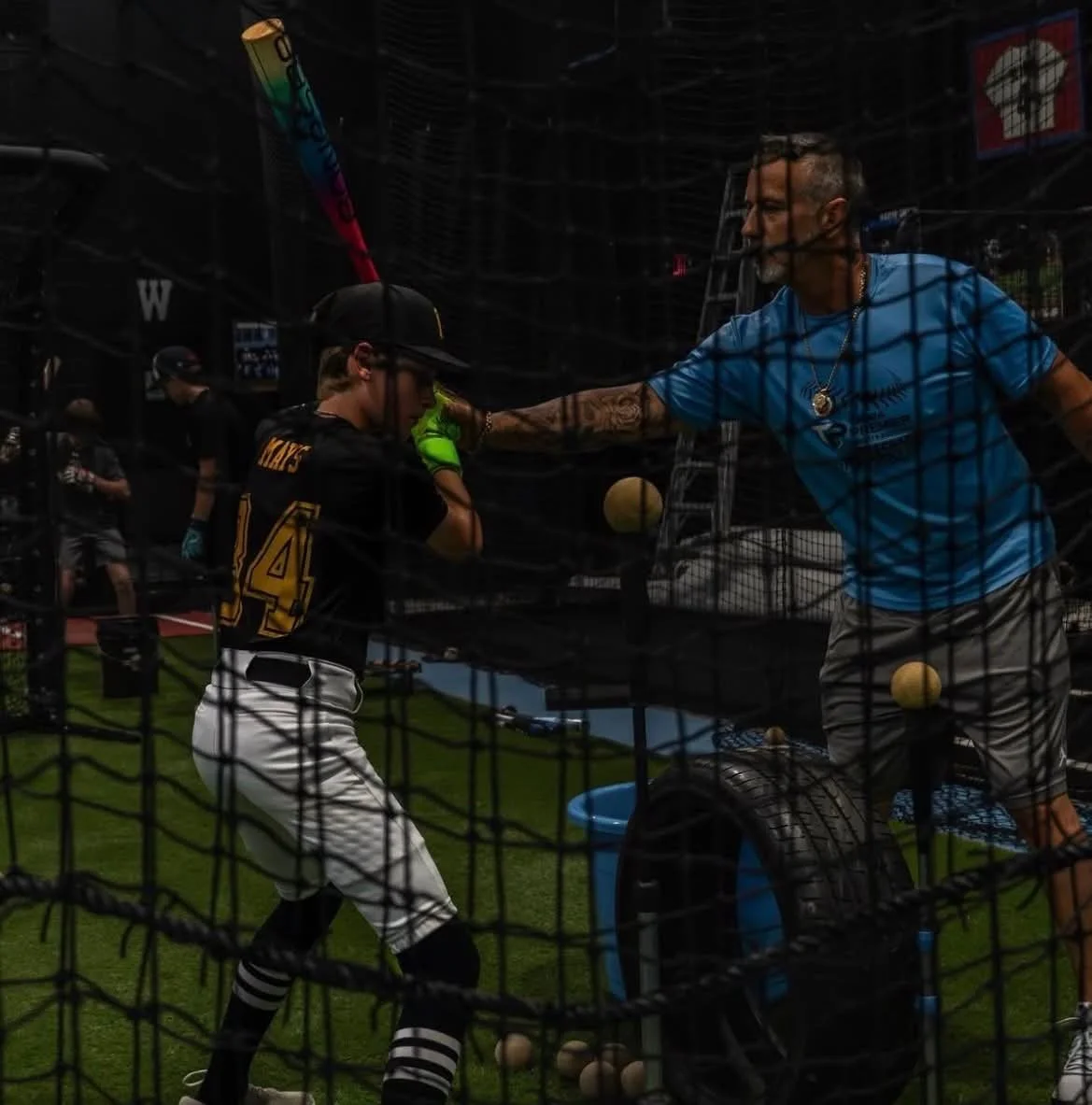 A young baseball player is at batting practice, leaning on a coach who is speaking to him through a batting cage. The environment is a baseball field with other people in the background.