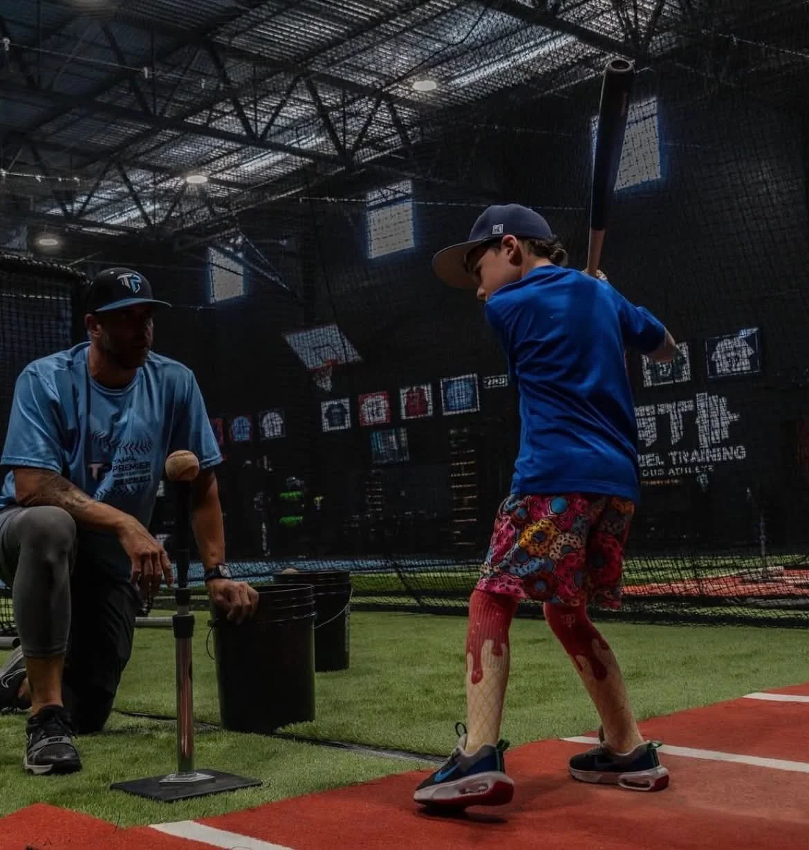 A young boy practicing baseball in a batting cage with a coach guiding him, inside an indoor sports facility.