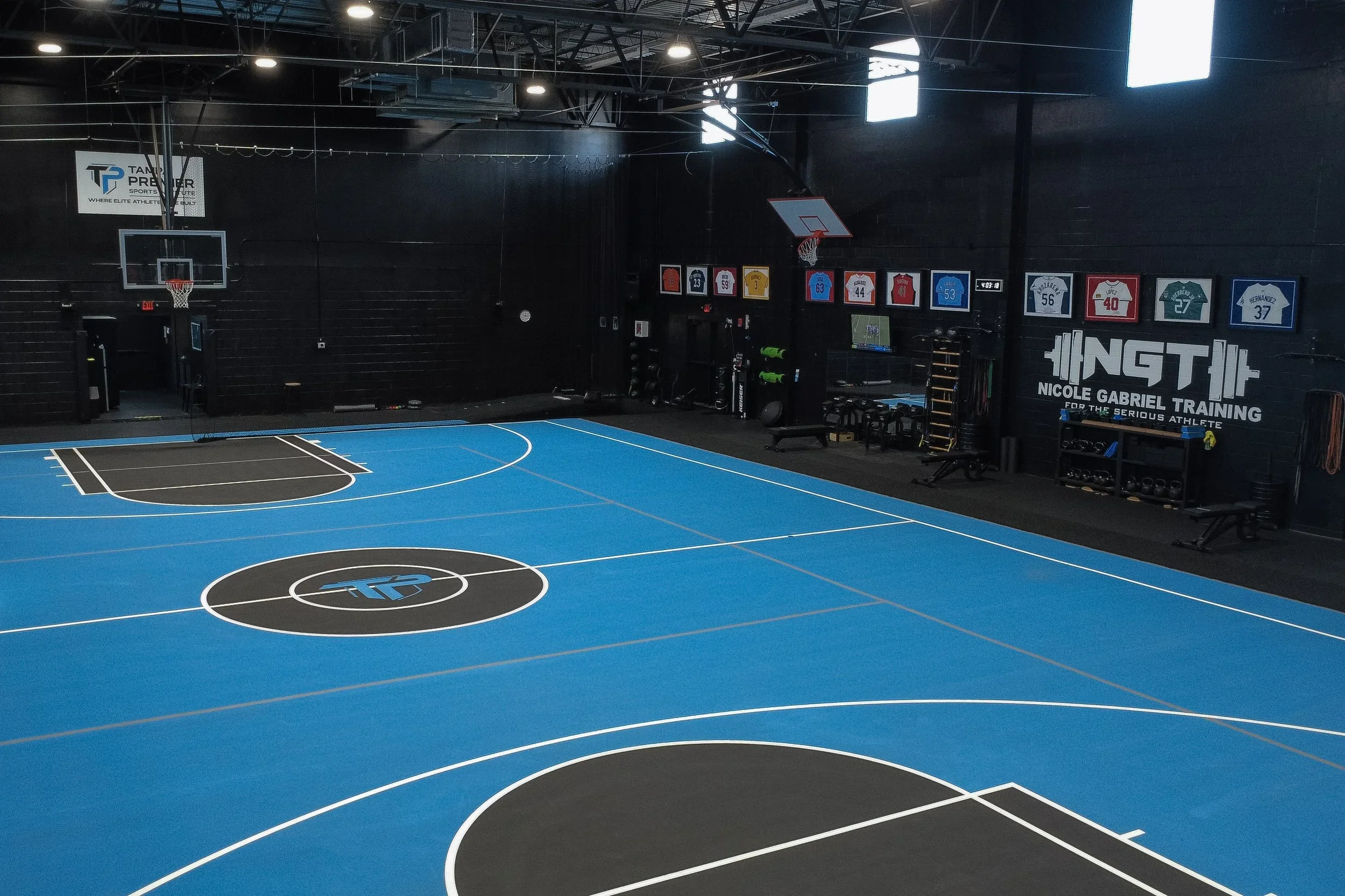 Indoor gym featuring a blue and black basketball court with white lines, two basketball hoops, a TV, and sports jerseys and training equipment on the walls.