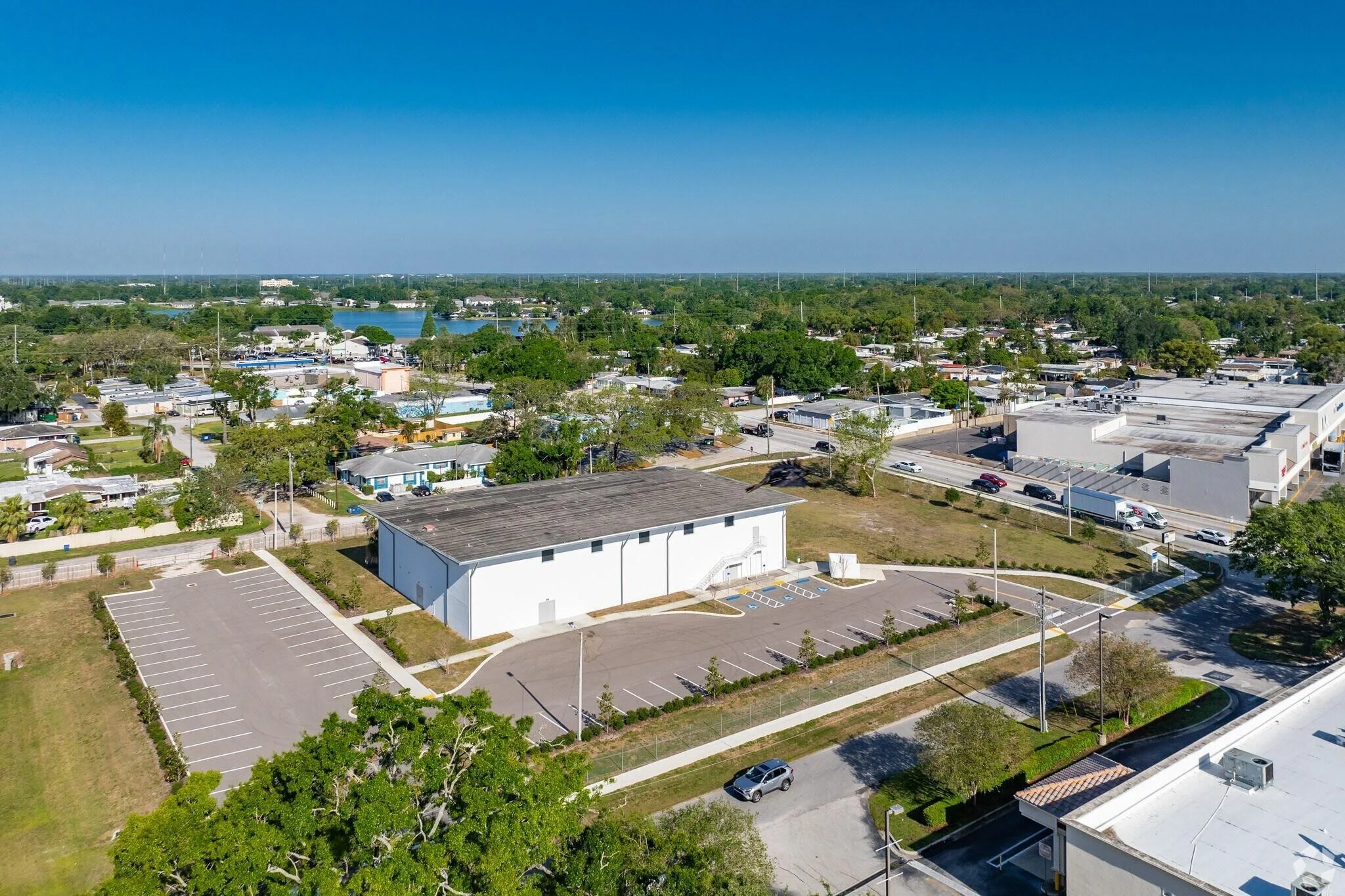Aerial view of a parking lot, with a white building in the foreground, surrounded by trees and roads, in a suburban area with many houses and commercial buildings, under a clear blue sky.