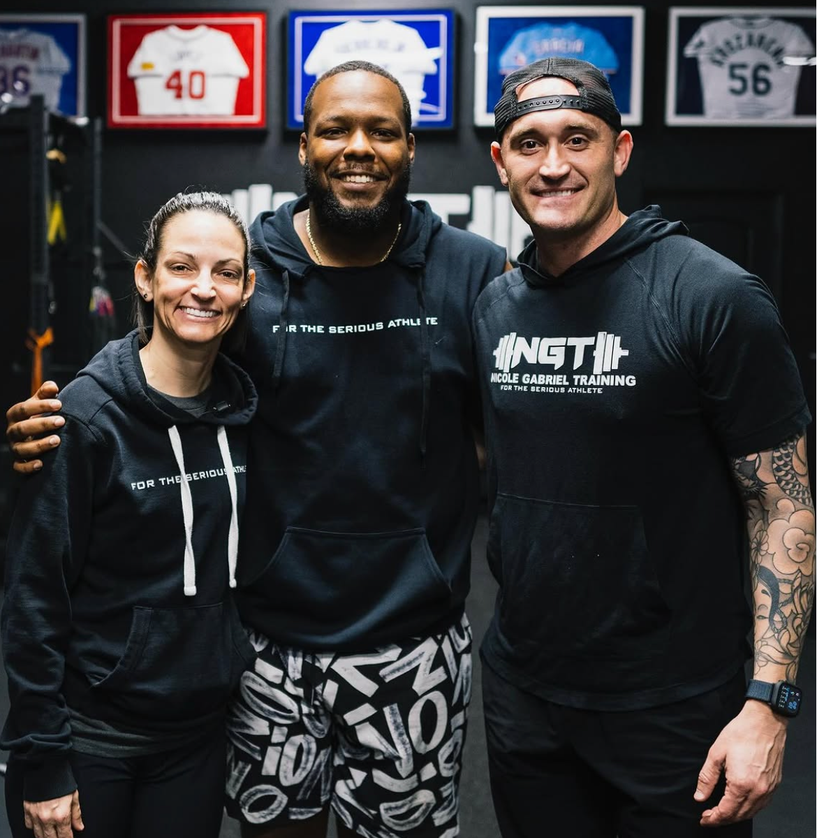 Three smiling people standing together in a gym, wearing black athletic hoodies with white text, with framed sports jerseys hanging on the wall behind them.