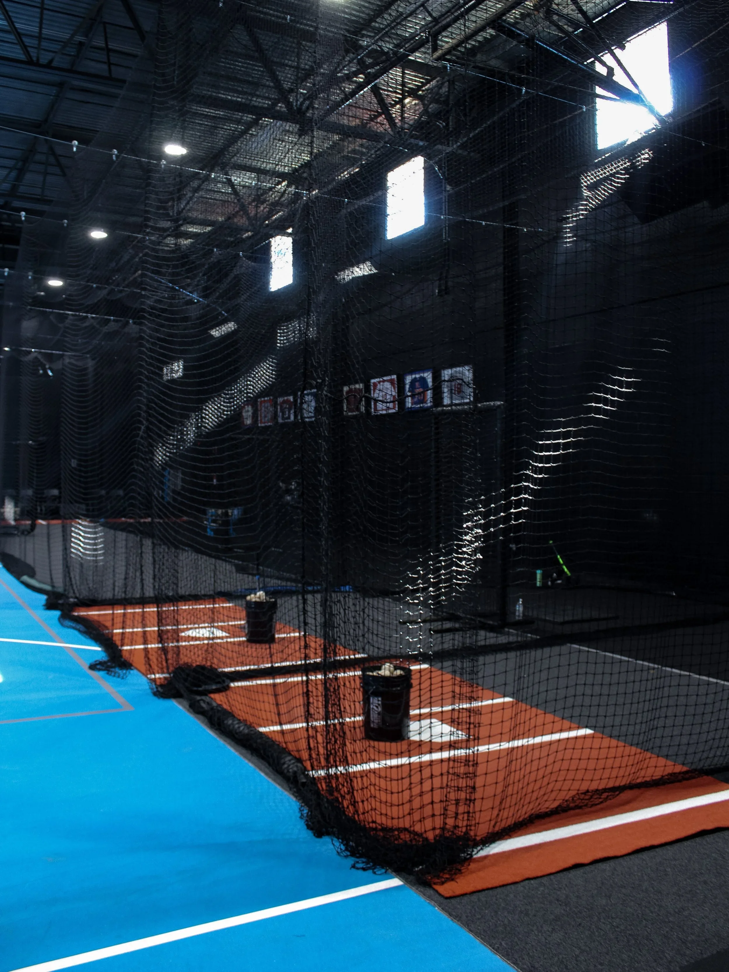 Indoor sports facility with batting cages, black nets, and blue flooring, illuminated by overhead lights and natural light from windows.