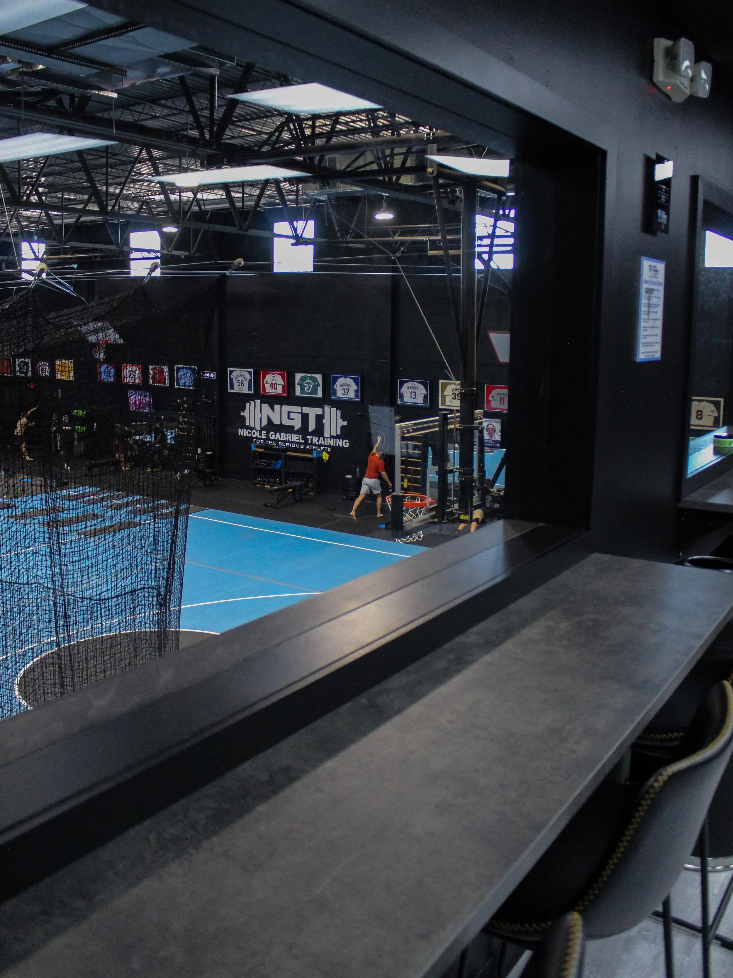 View through a window of an indoor sports training facility with a blue court, fitness equipment, and banners on the wall, with someone in a red shirt preparing for training.