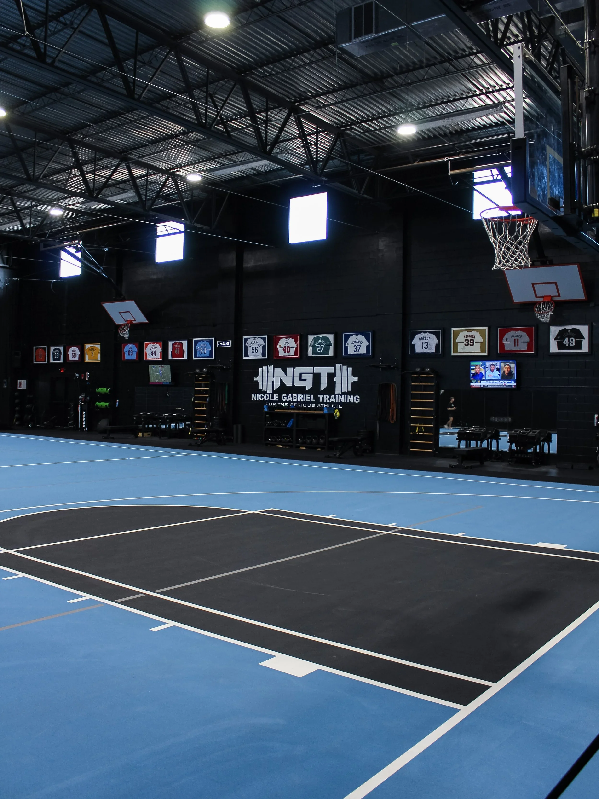 Empty blue and black indoor basketball court with basketball hoop, scoreboards, and mounted televisions on black walls, and banners hanging from ceiling.