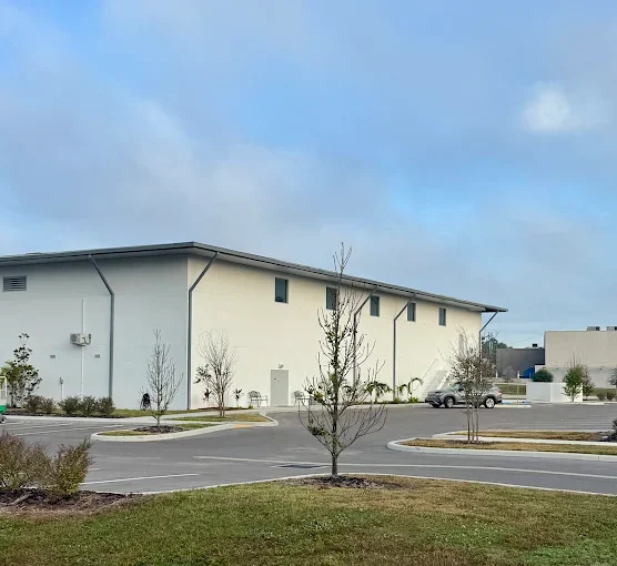 Empty parking lot in front of a large, light-colored commercial building with small trees and grassy areas