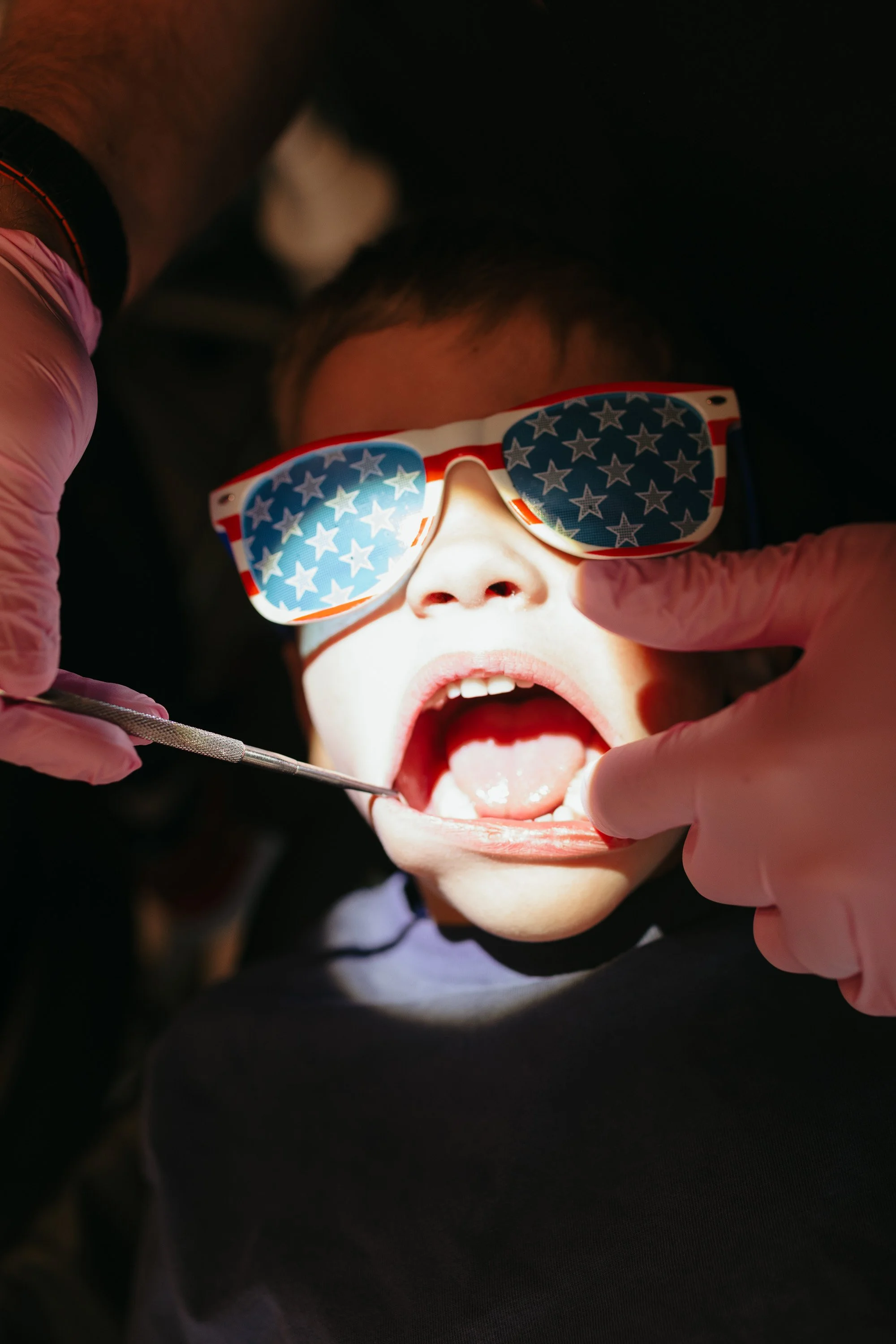 Child at the dentist with American flag sunglasses, open mouth, being examined with a dental tool.