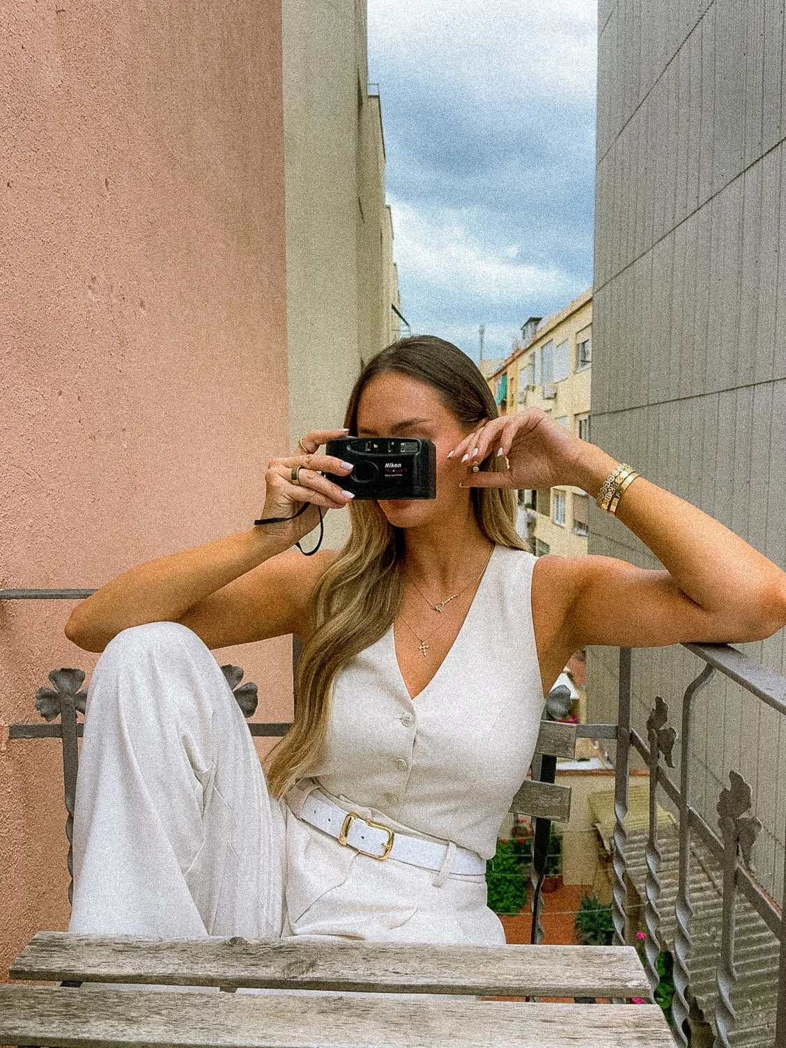 A woman sitting on a balcony taking a photo with a camera, wearing white pants and a sleeveless white top, with a grey and peach wall in the background.
