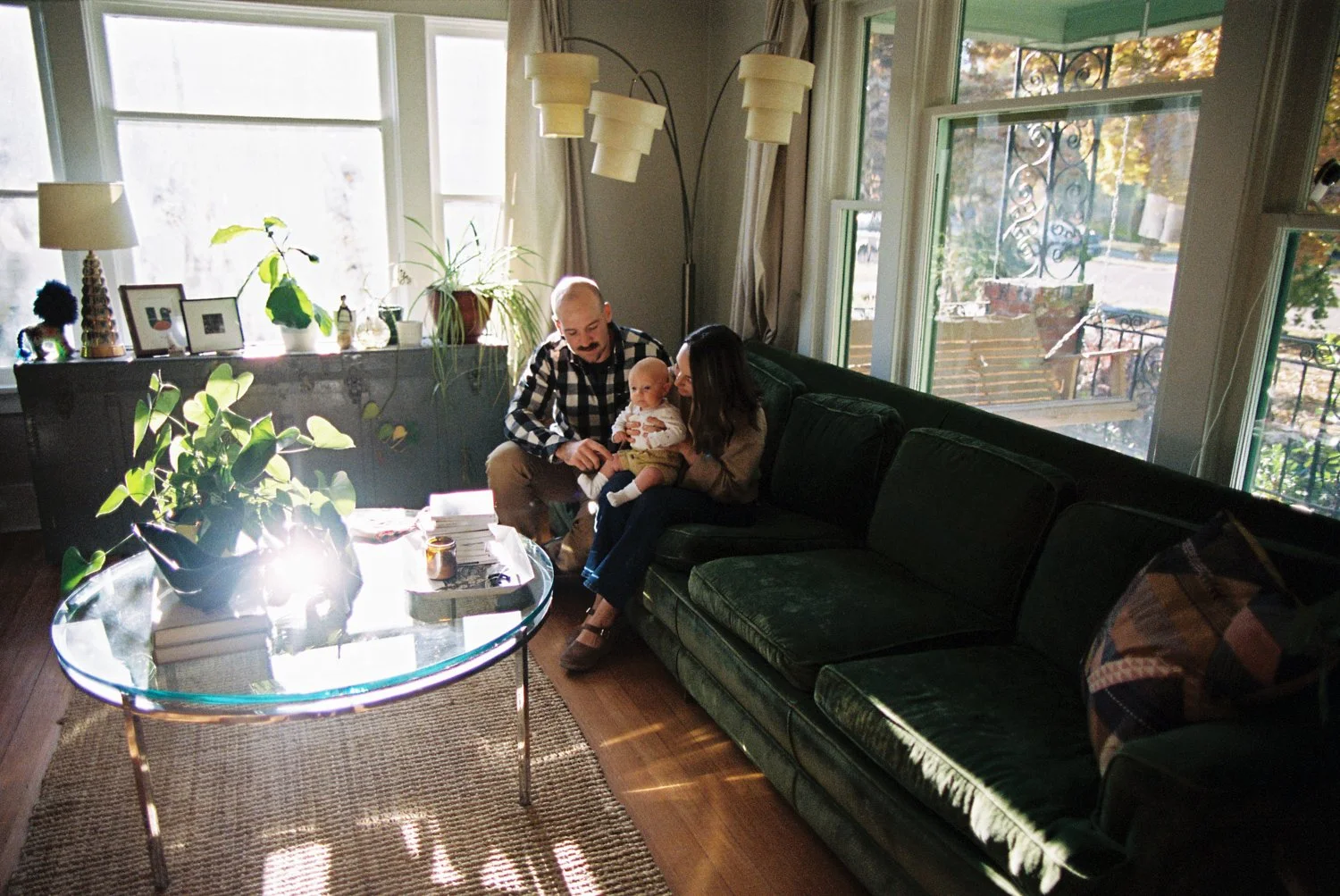A family sitting on a dark green couch in a sunlit living room, with two adults and a baby.