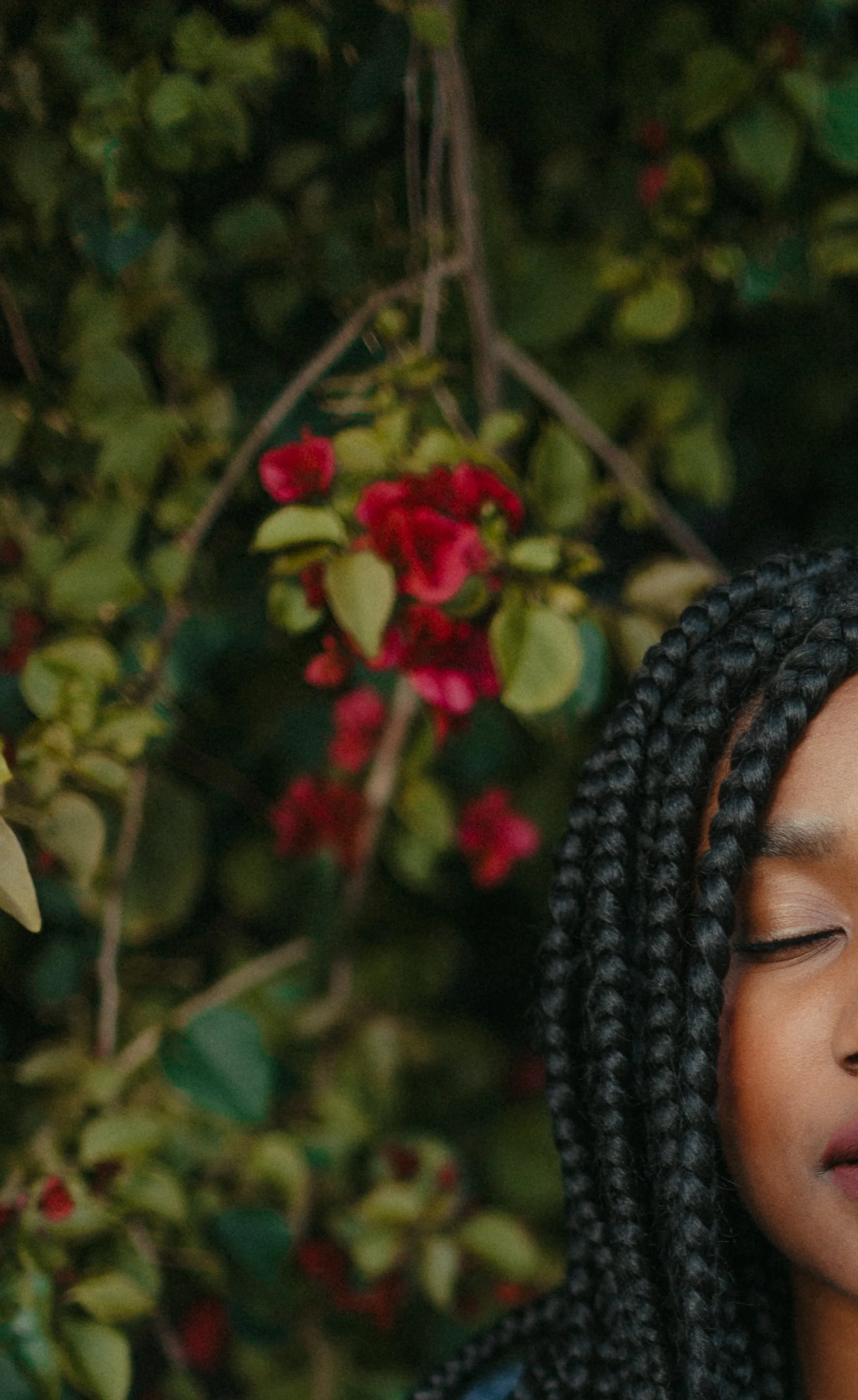 Part of a woman with closed eyes and long braided hair in front of green foliage and pink flowers.