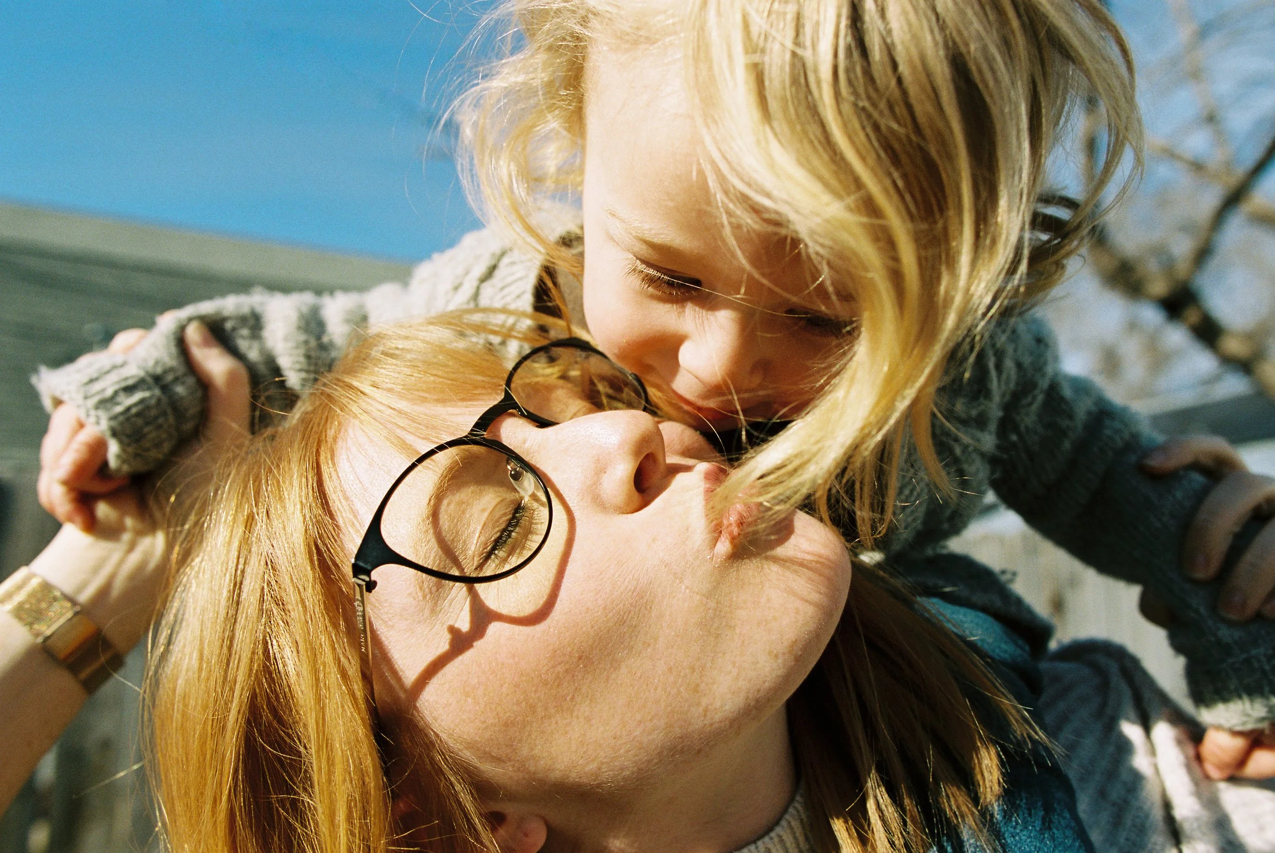 A woman with red hair and glasses kissing a young girl with blonde hair and a gray sweater, outdoors on a sunny day in Boise, Idaho.