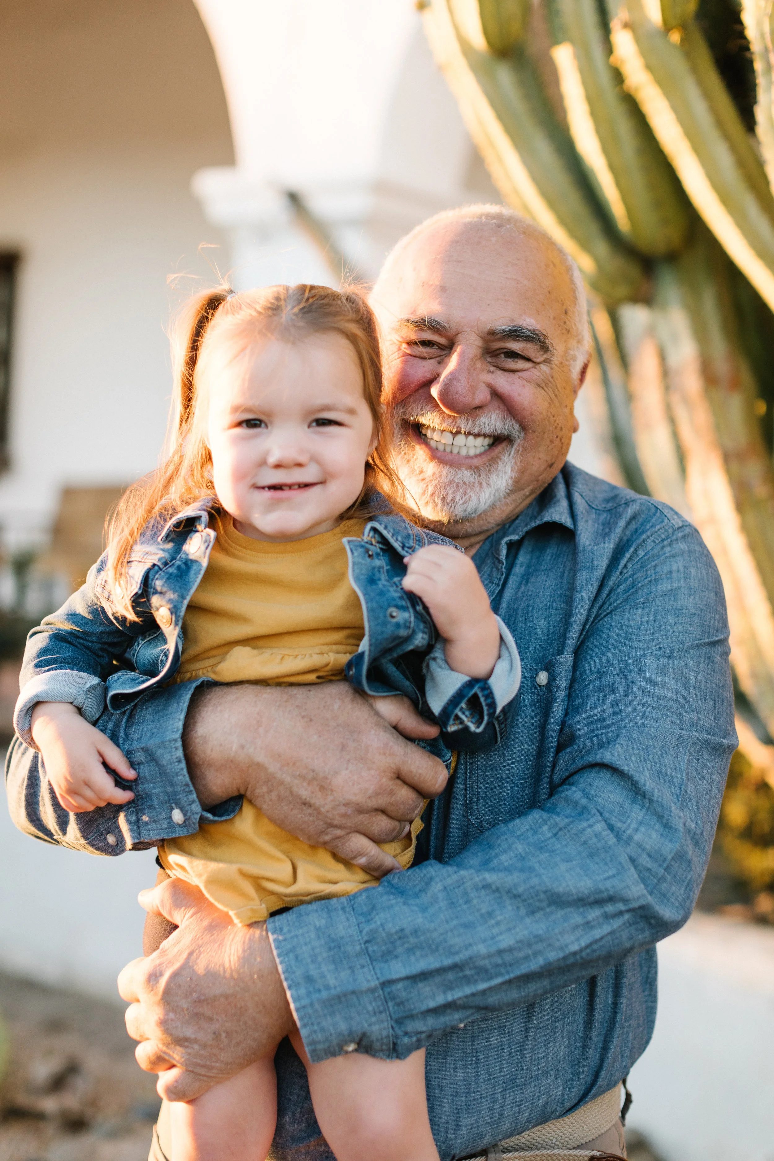 An elderly man with a white beard and a young girl with long hair smiling while being held outdoors near a cactus in warm sunlight.