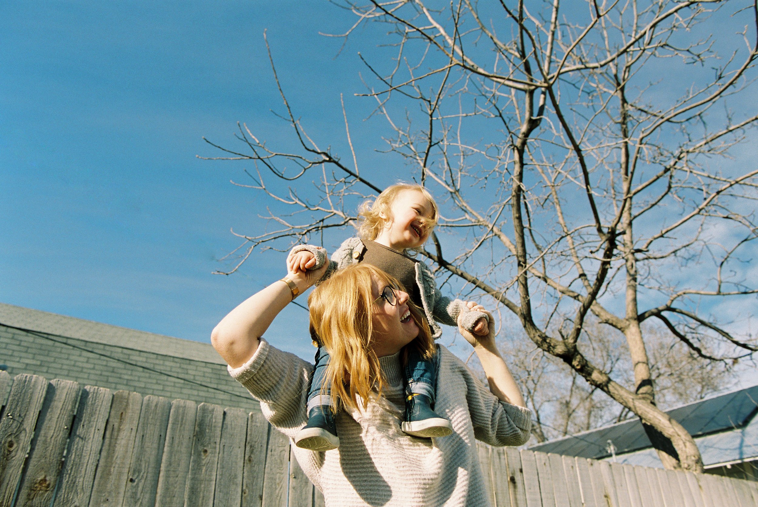 A woman with red hair and glasses carrying a happy child on her shoulders, playing outdoors on a sunny day with a clear blue sky and bare trees in the background.