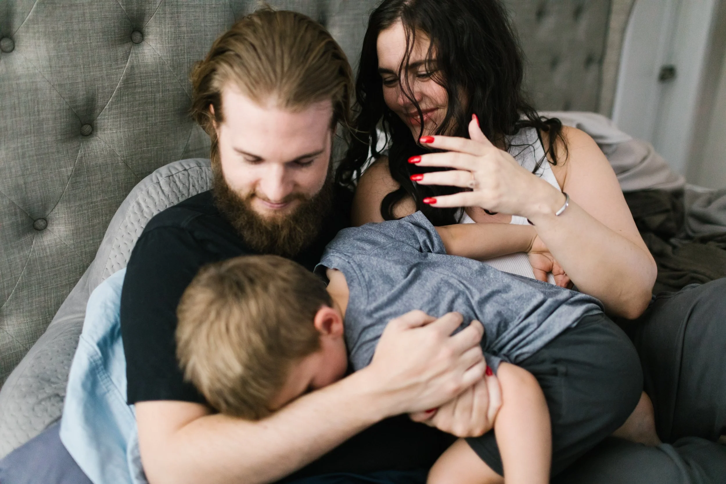 A family of four, including two children, relax on a bed. The mother has dark, curly hair and red nails, and is smiling as she watches her children. The father, with a beard and long hair, is holding and comforting one of the children who is lying on