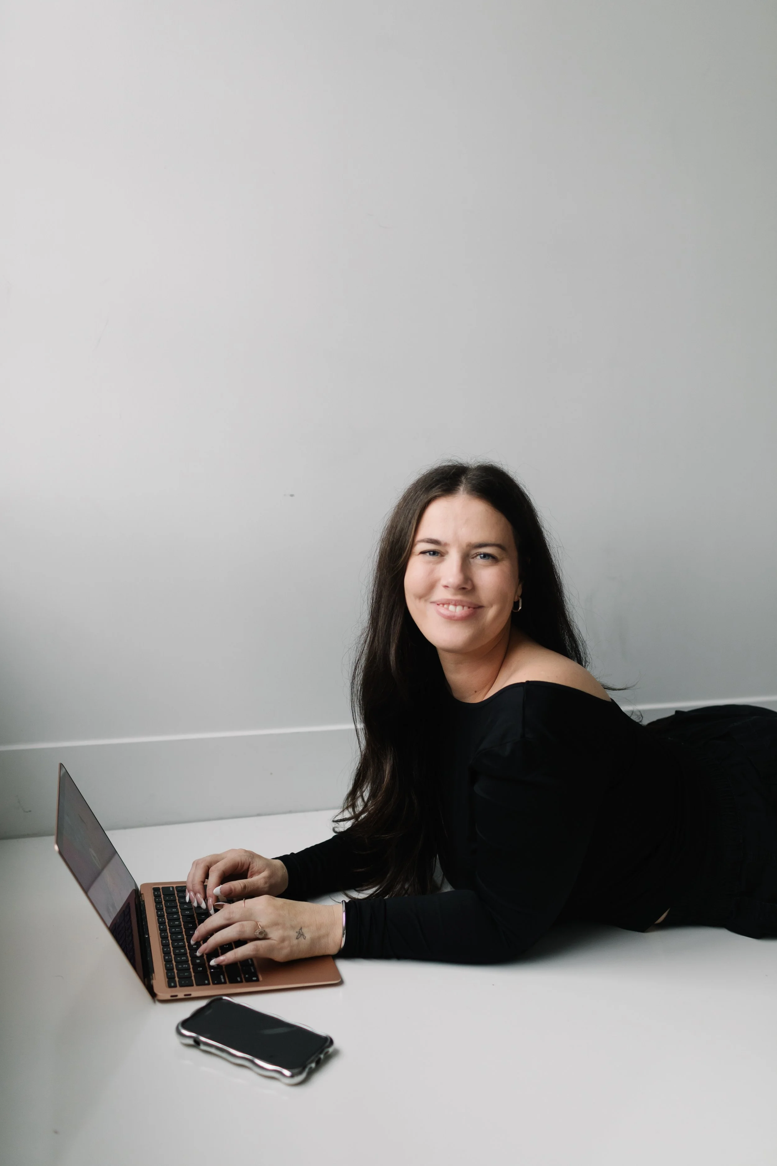 A woman with long dark hair lying on her stomach on a white table, using a laptop, with a smartphone beside her, against a plain light grey wall.
