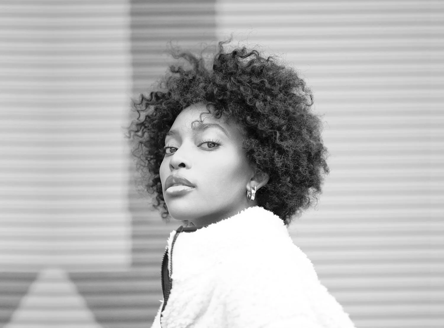 A black and white portrait of a woman with curly hair, wearing earrings and a cozy white sweater, looking over her shoulder.