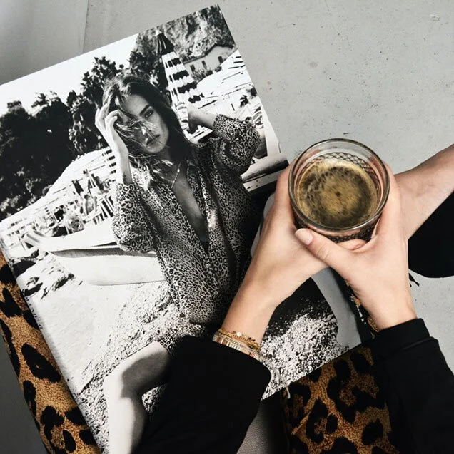 A person holding a glass of coffee or tea over an open black-and-white magazine featuring a woman wearing sunglasses and a patterned dress at a beachside location.