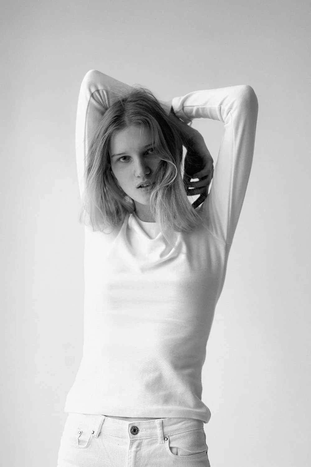 Black and white photo of a young woman with shoulder-length hair, wearing a long-sleeved shirt and light-colored pants, posing with her hands behind her head against a plain background.