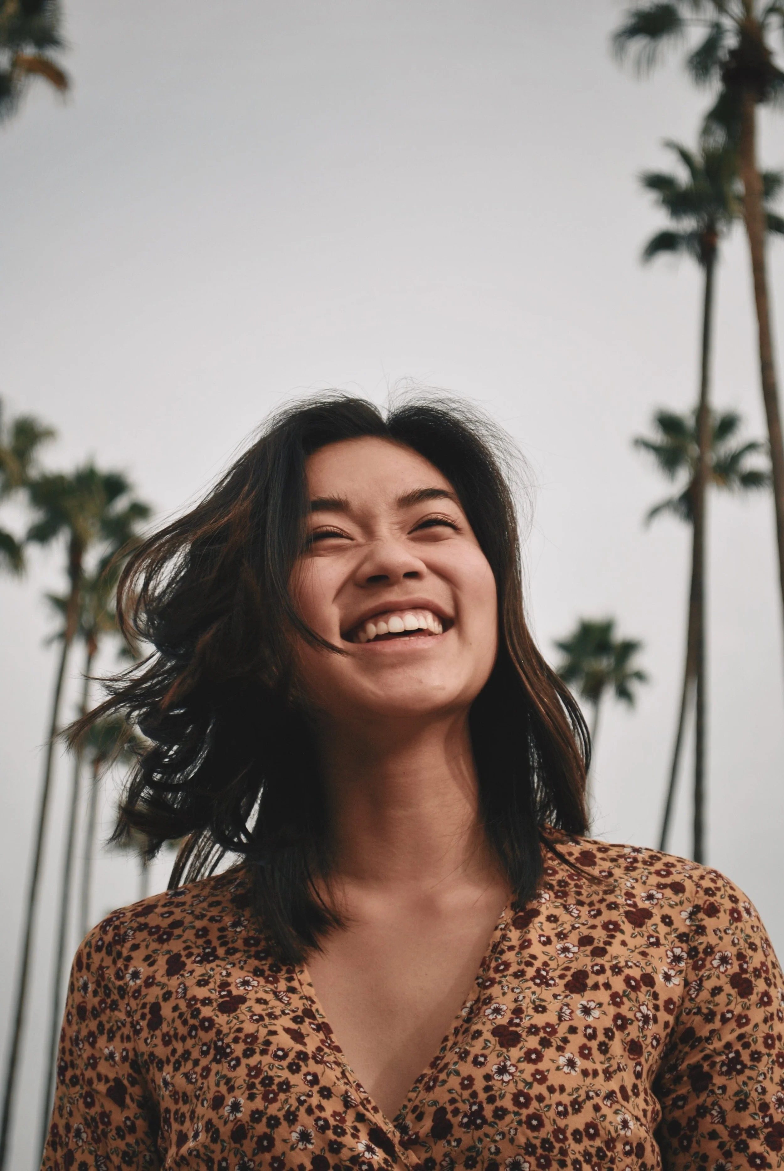 A woman with shoulder-length dark hair smiling and looking up outdoors, with palm trees and a cloudy sky in the background.