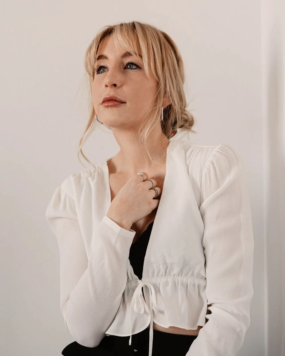 A young woman with blonde hair styled in a loose bun, wearing a white blouse and black top, standing against a plain background with a thoughtful expression.