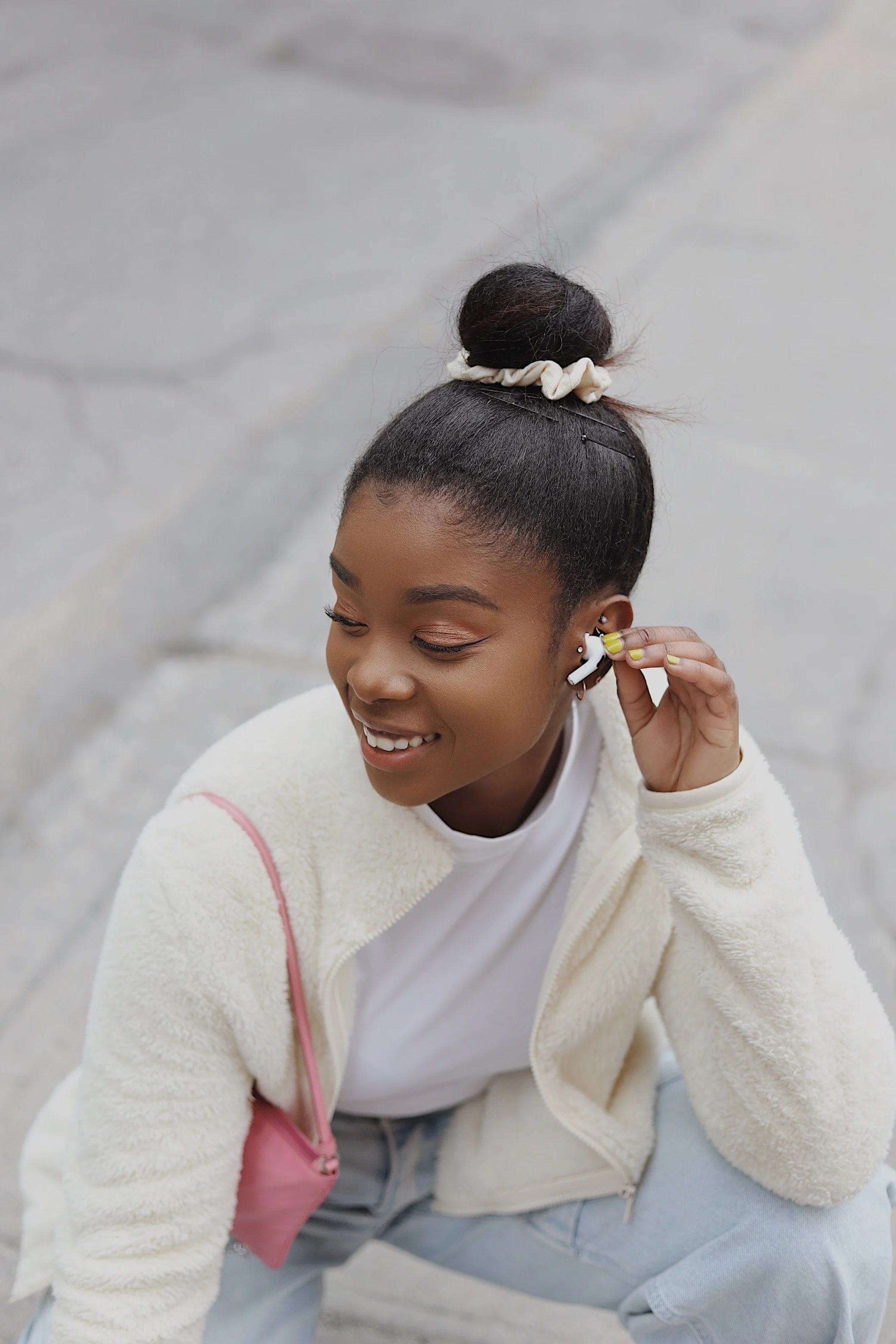 A young woman with dark hair in a bun, wearing a white fleece jacket and light-colored jeans, smiling and adjusting her AirPods while sitting outdoors on a concrete surface.