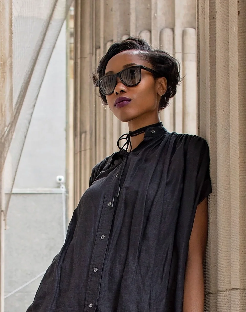 A woman with short, dark hair wearing large black sunglasses, a black choker, and a loose-fitting black shirt, standing against a stone architectural background.