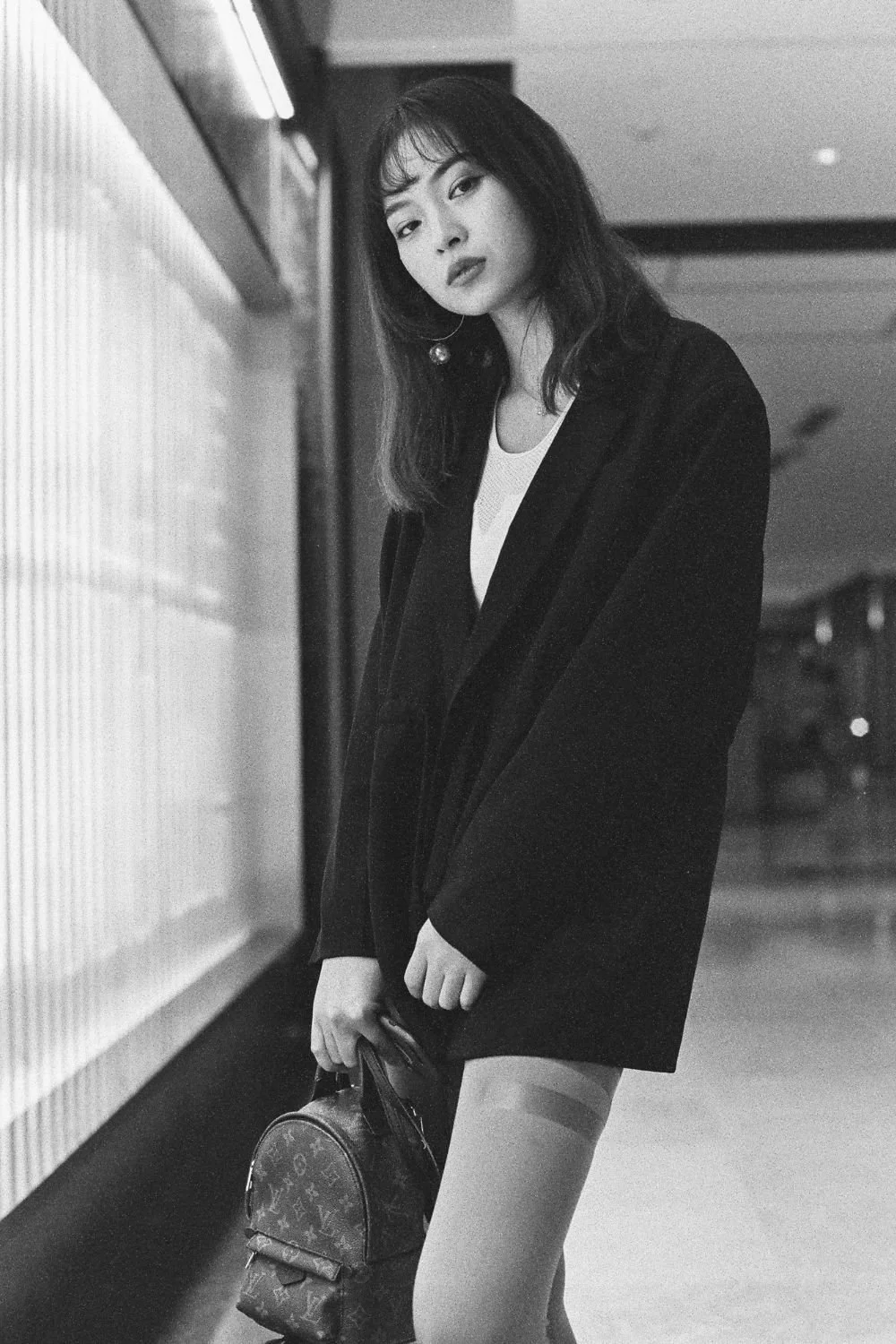 Black and white photo of a young woman with long hair, wearing a blazer over a top, holding a small Louis Vuitton handbag, standing in an indoor setting with a contemplative expression.