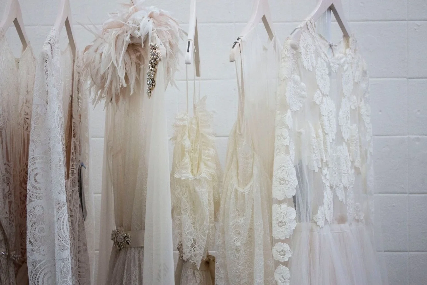 Collection of white lace and fabric dresses hanging on a rod, with decorative accessories like feathers and jewelry, in front of a white brick wall.