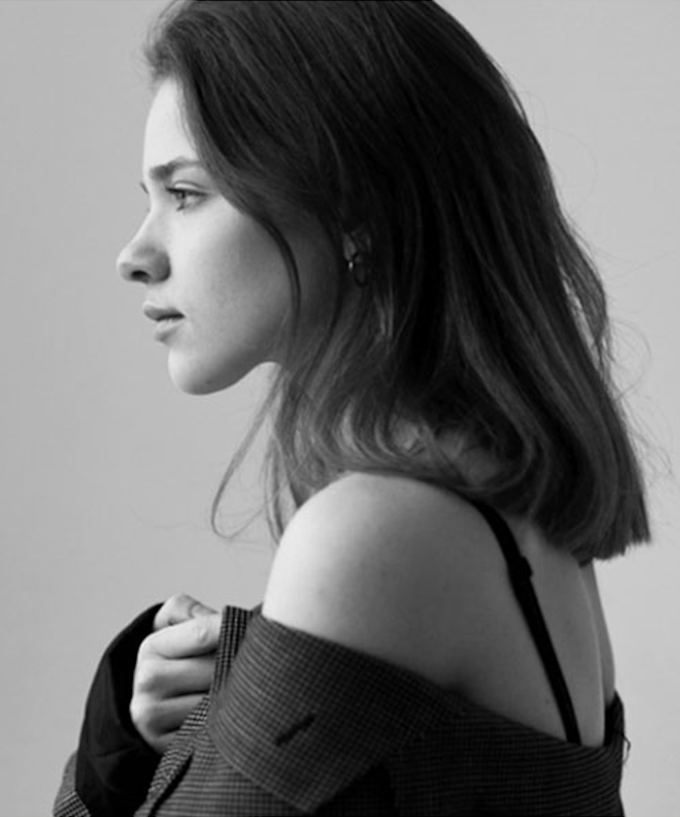 A black and white photograph of a woman in profile with shoulder-length dark hair, wearing a checkered jacket slipping off her shoulder, revealing a black strap underneath, holding the jacket with one hand.