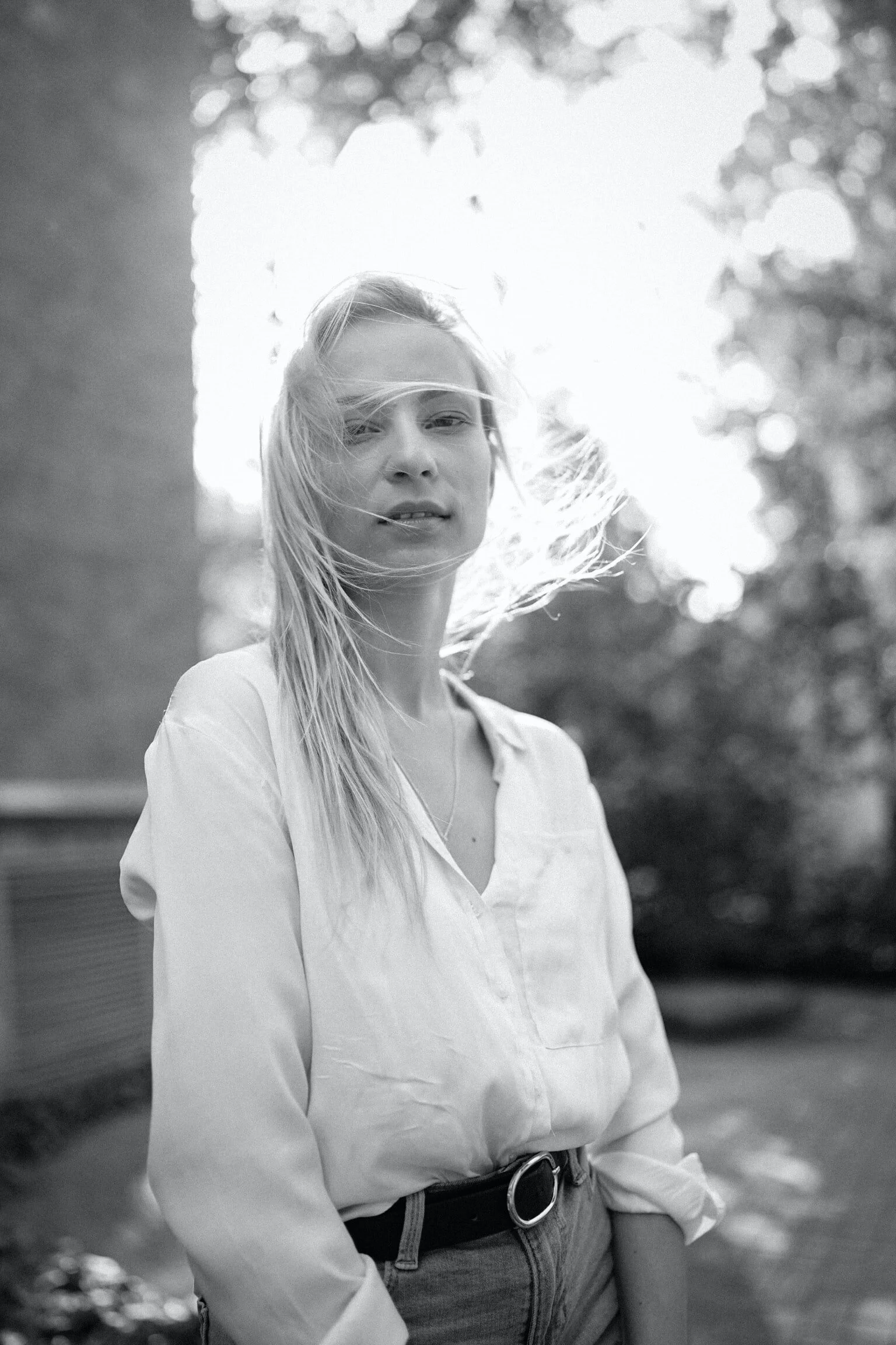 A woman with long hair standing outdoors, dressed in a light-colored shirt and jeans, with wind blowing her hair.