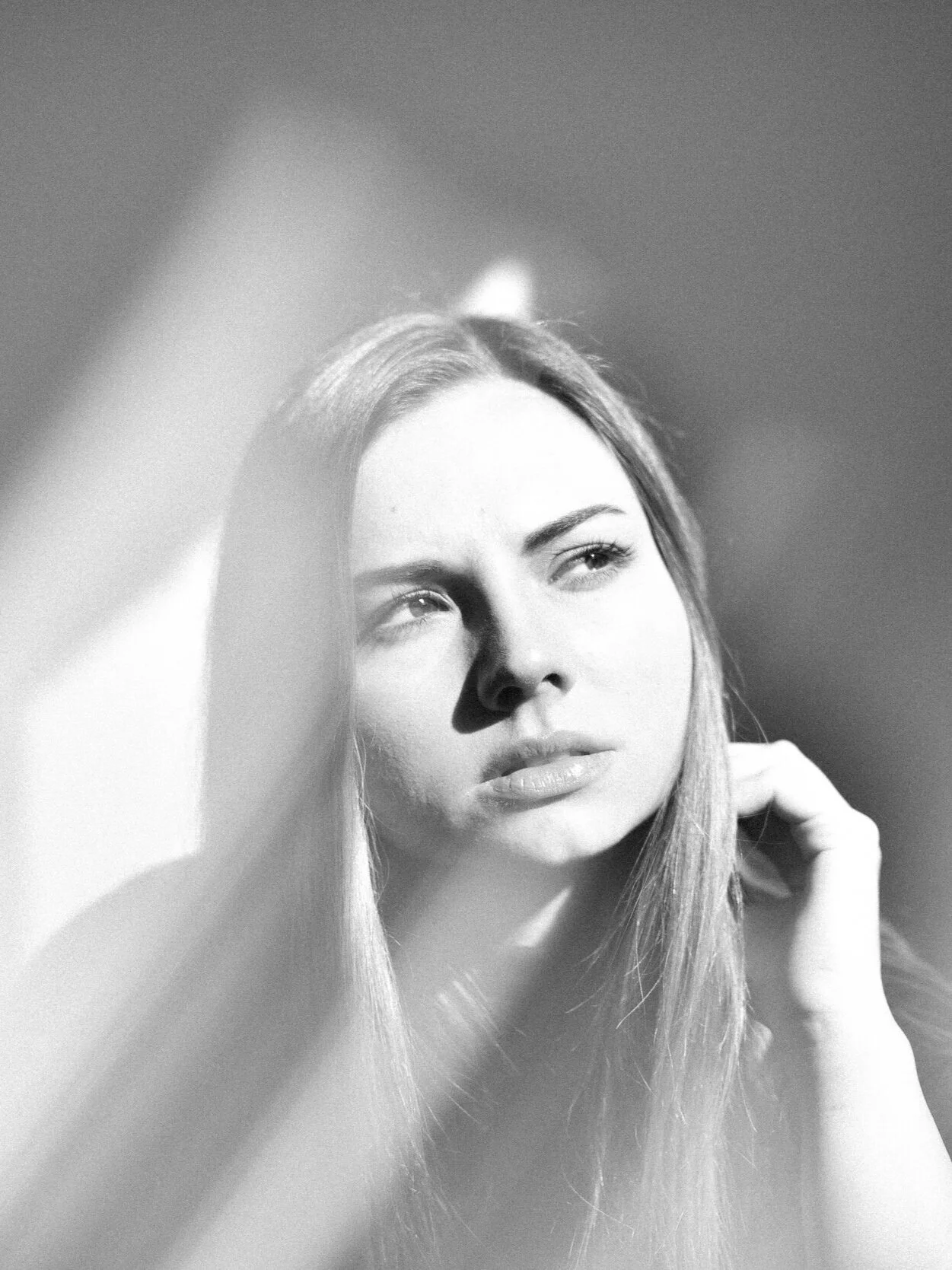 Black and white portrait of a woman with long hair, her face partly shadowed, looking thoughtfully to the side with a hand touching her neck.
