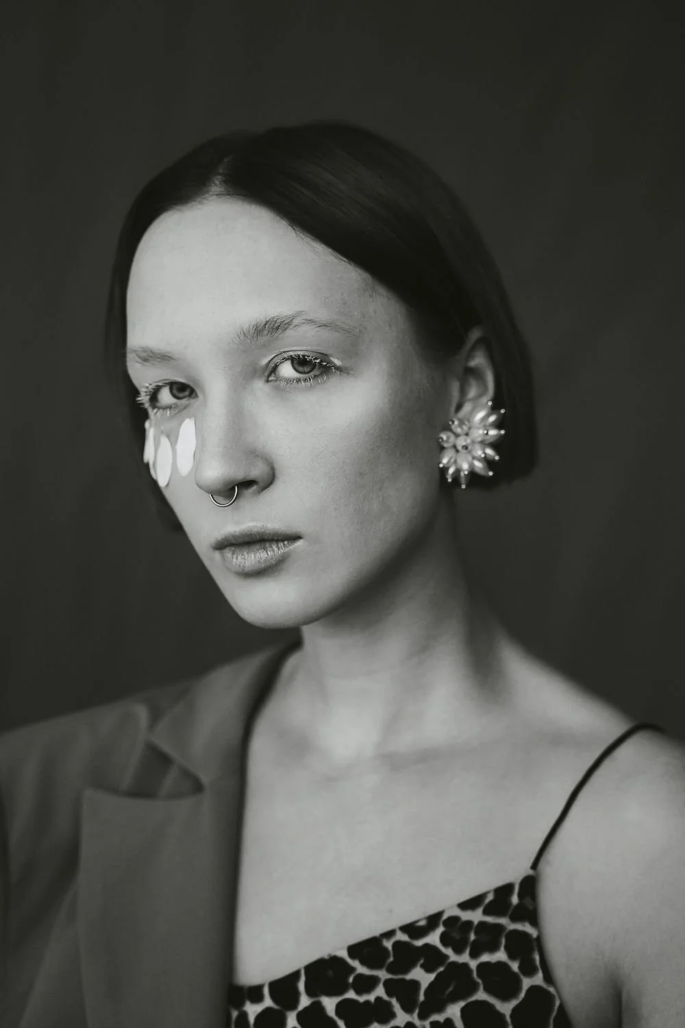 Black and white portrait of a woman with short dark hair, wearing floral earrings and a leopard print top, with tears on her cheek.