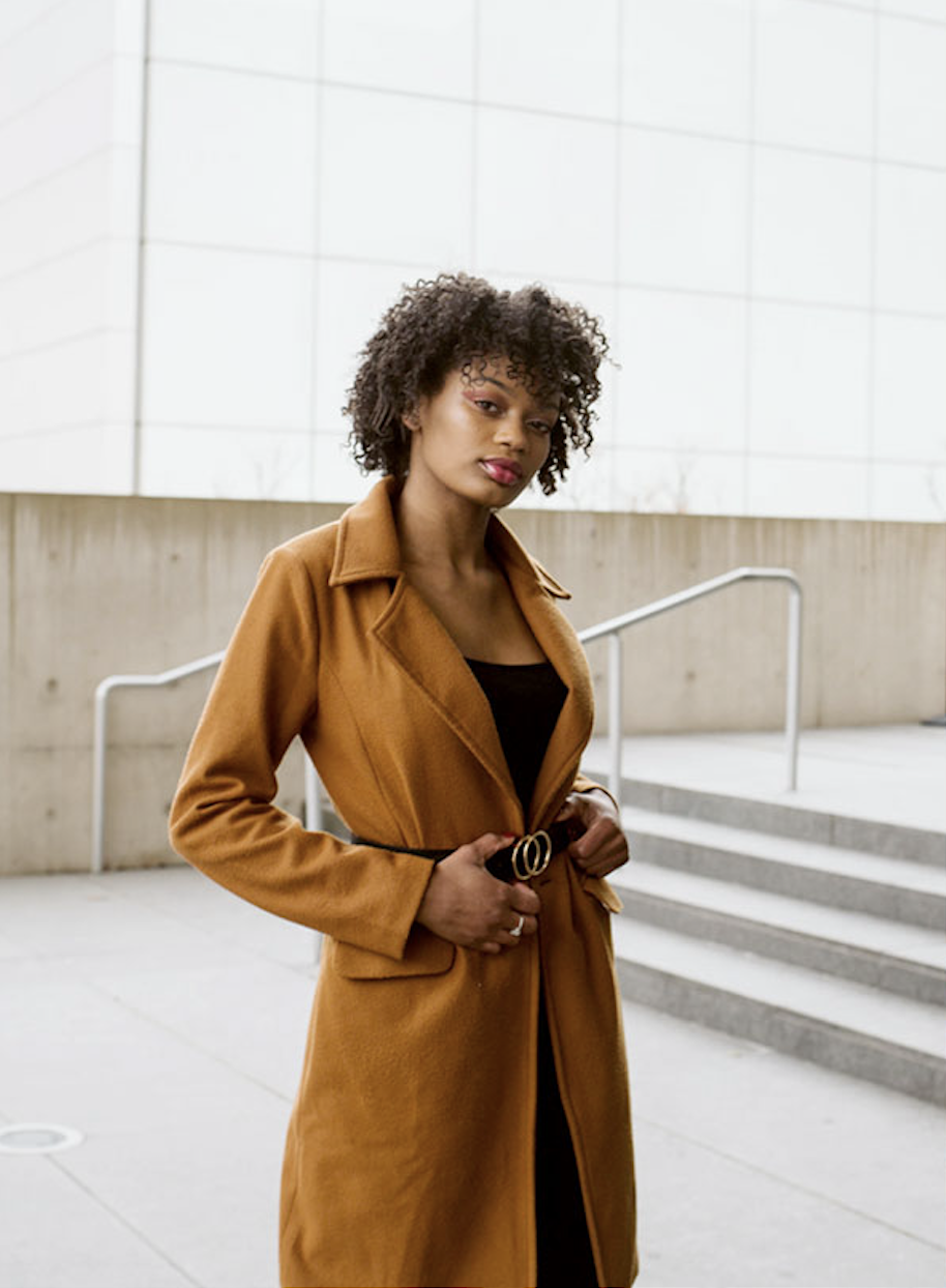 A woman with curly hair wearing a brown coat and black top standing outdoors near steps in an urban setting.