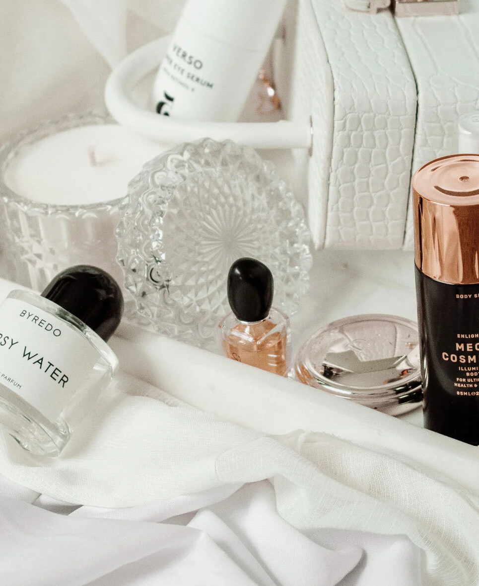 A collection of skincare and cosmetic products, including perfume bottles, a candle, a mirror, and a decorative crystal dish, arranged on a white surface with white fabric.