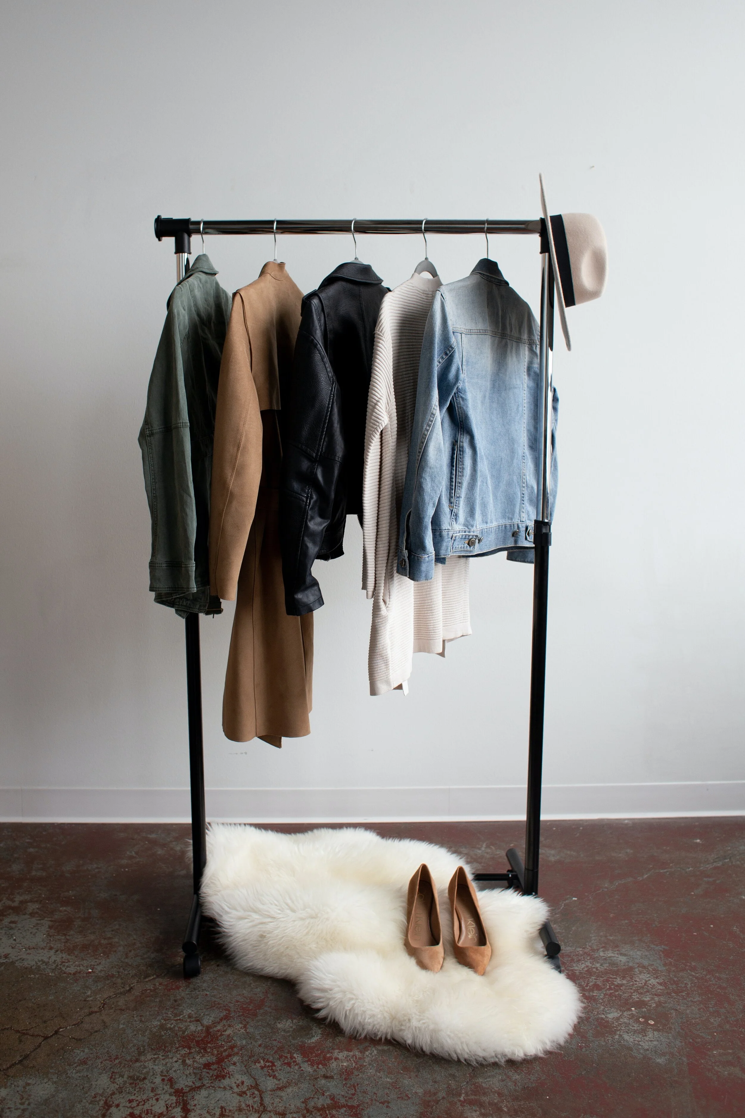 Clothing rack with five articles of clothing, a hat hanging on the side, and a pair of tan shoes on a white fluffy rug in front of the rack.