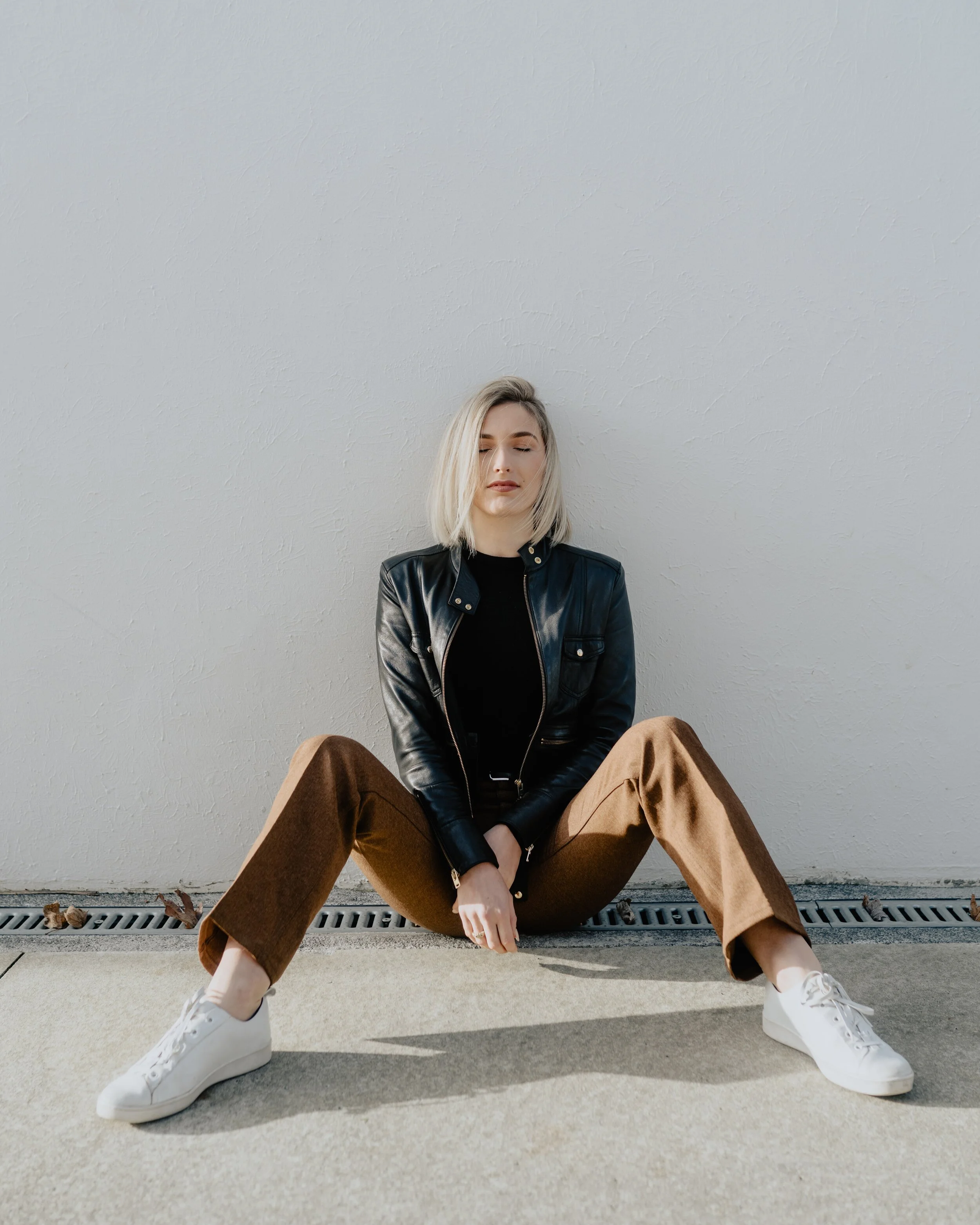 Woman sitting on the ground against a white wall, wearing a black leather jacket, black top, brown pants, and white sneakers with eyes closed.