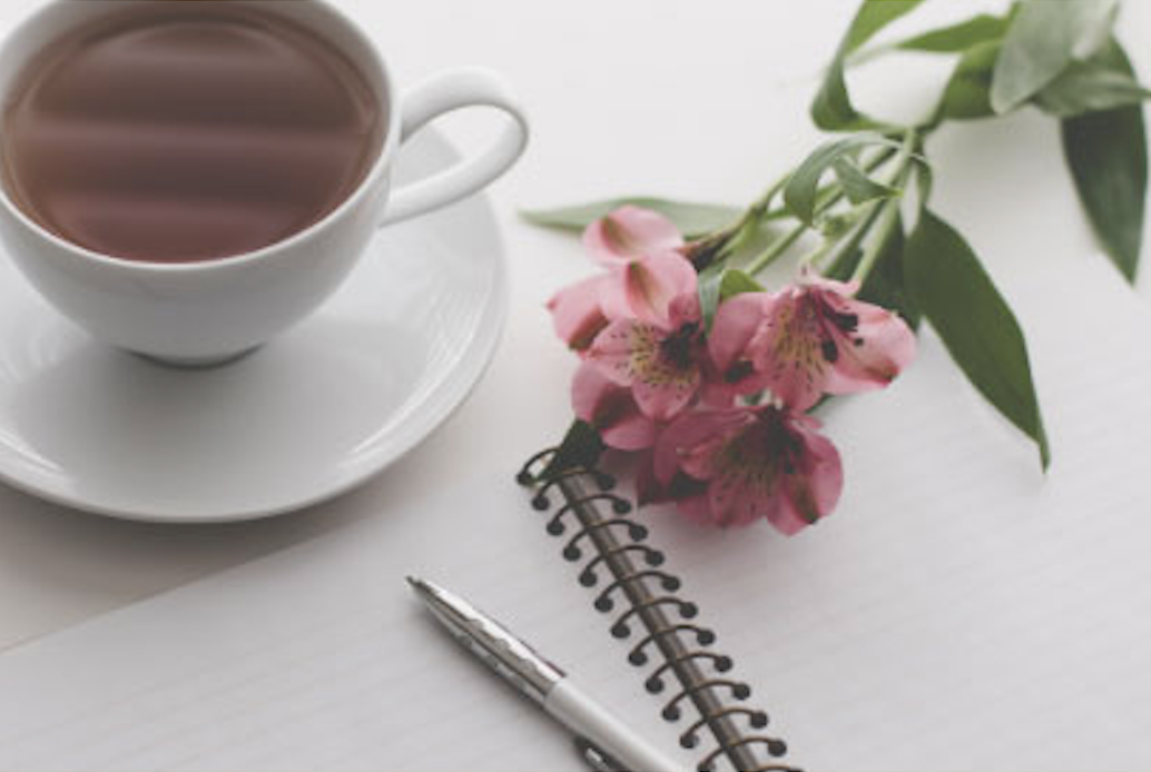 A white teacup filled with tea on a matching saucer, pink flowers with green leaves, an open notebook, and a silver pen on a white surface.