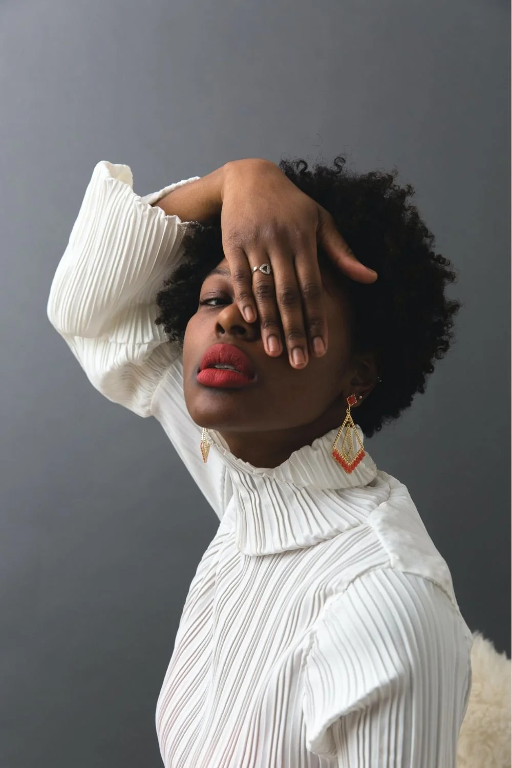 A woman with curly black hair wearing a white pleated high-neck top, red earrings, and red lipstick posing with her hand covering one eye against a plain gray background.