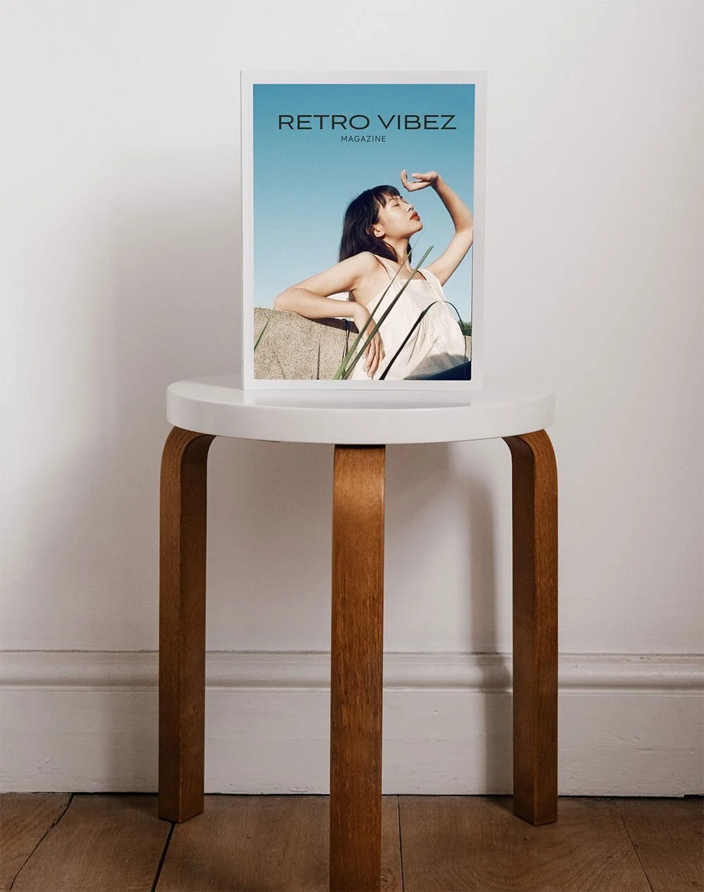 A magazine titled 'RETRO VIBEZ' with a woman on the cover, sitting outdoors against a stone wall, wearing a white dress and looking upward, is displayed on a small round white and wooden table.