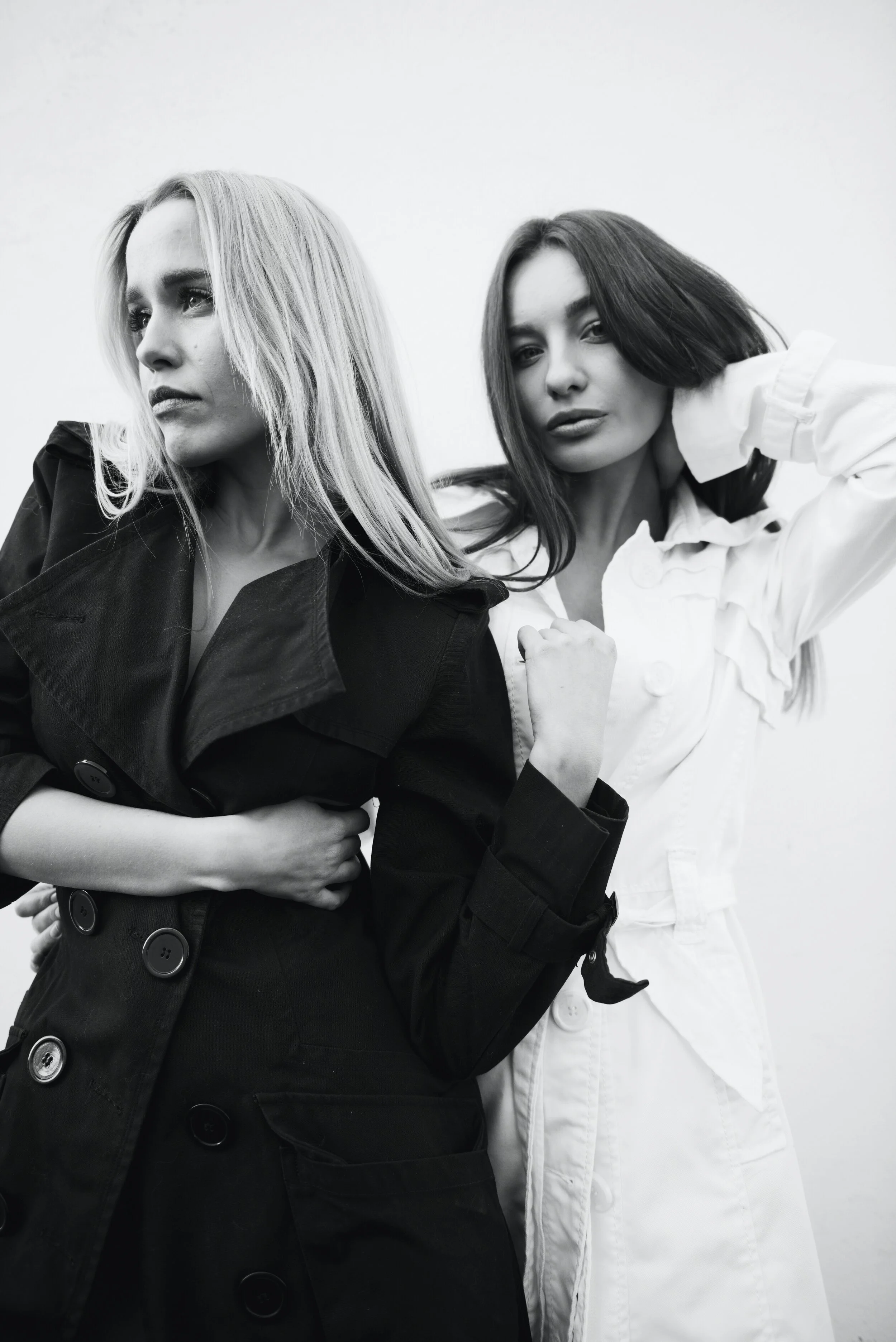 Two women with long hair, one in a black trench coat and the other in a white jacket, posing against a plain background.