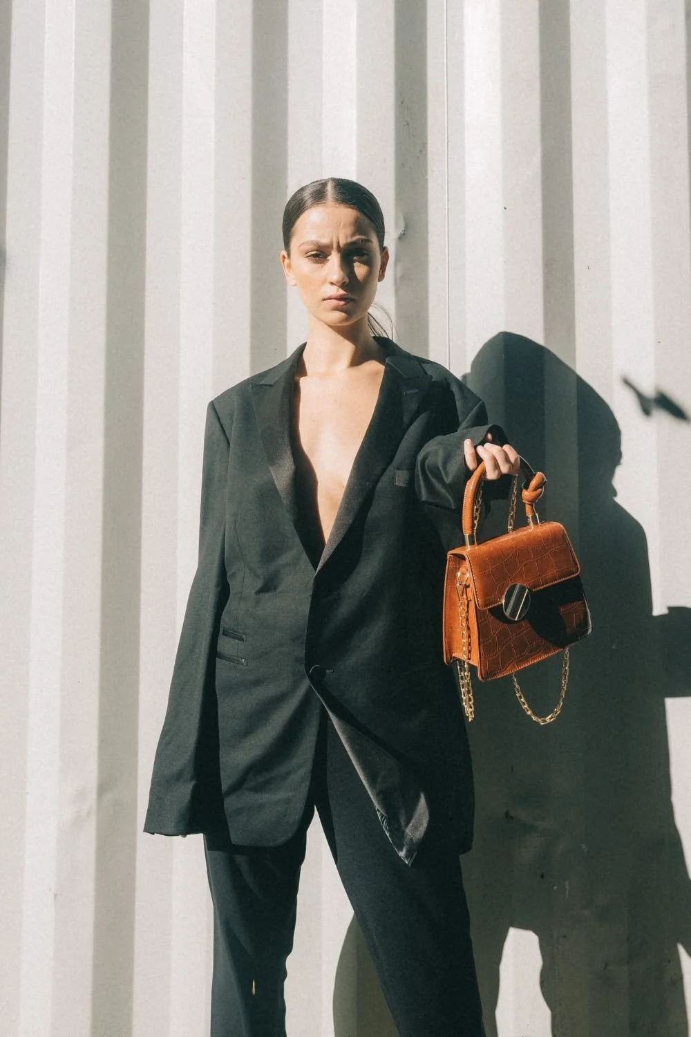 Woman in a black suit holding a brown handbag, standing against a striped white wall with shadows.